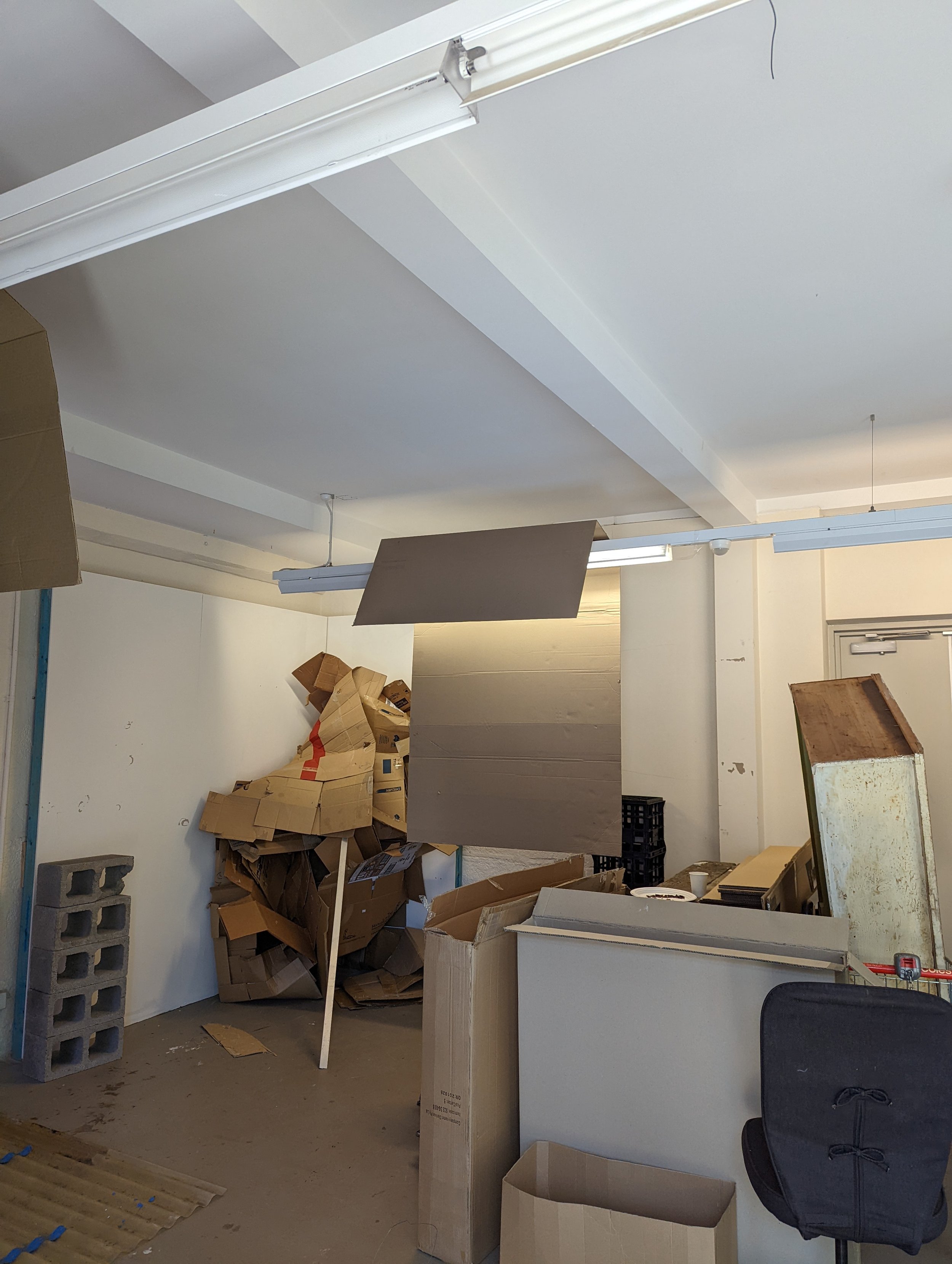 A cluttered room with numerous cardboard boxes, some stacked haphazardly, and various objects including a black plastic crate, a gray cinder block, and wooden panels. The ceiling has fluorescent lights, and there is a white partition or counter, with