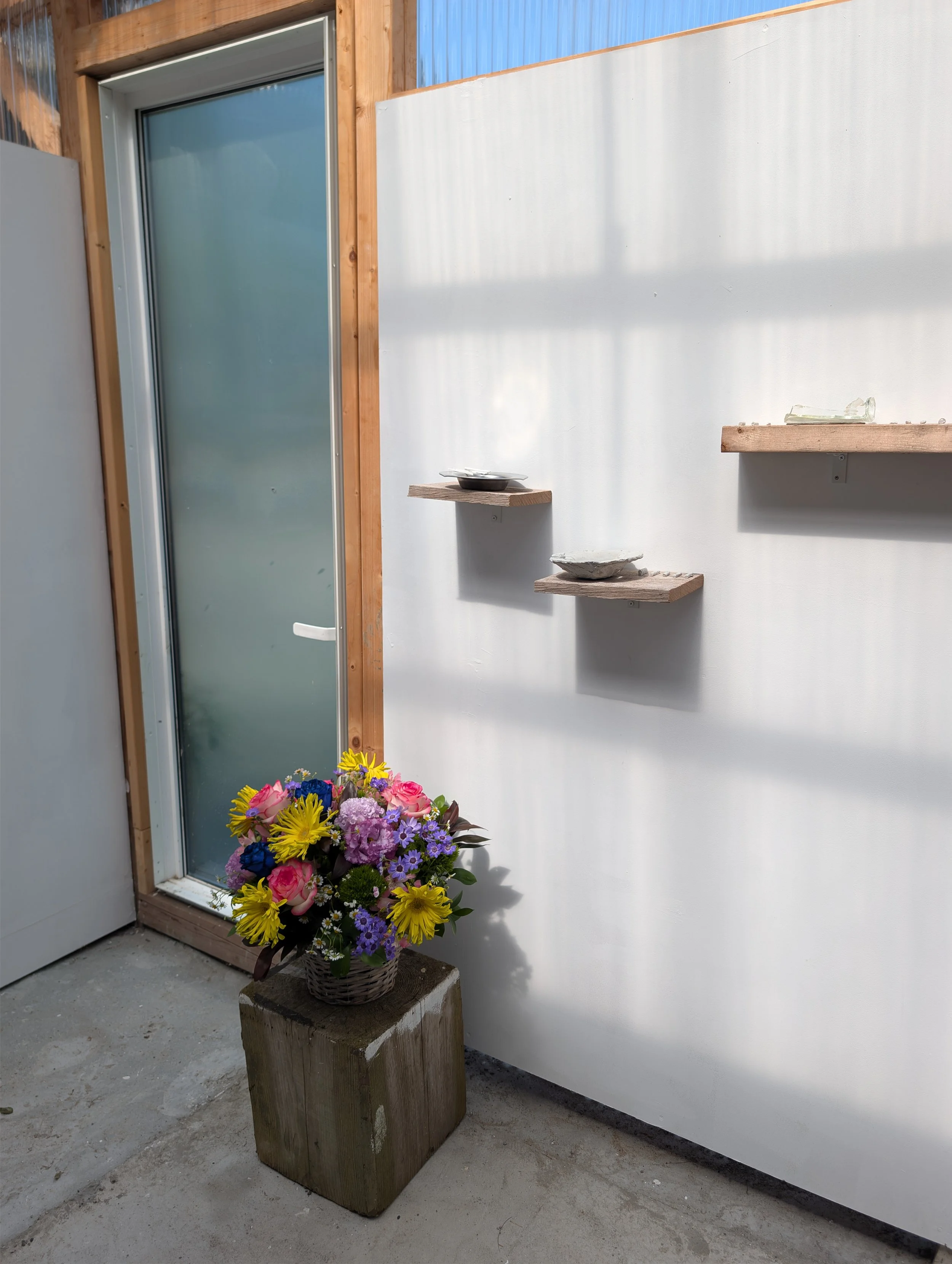A flower arrangement with colorful flowers in a basket, placed on a wooden block outside near a modern building with a sliding glass door and wooden and metal siding.