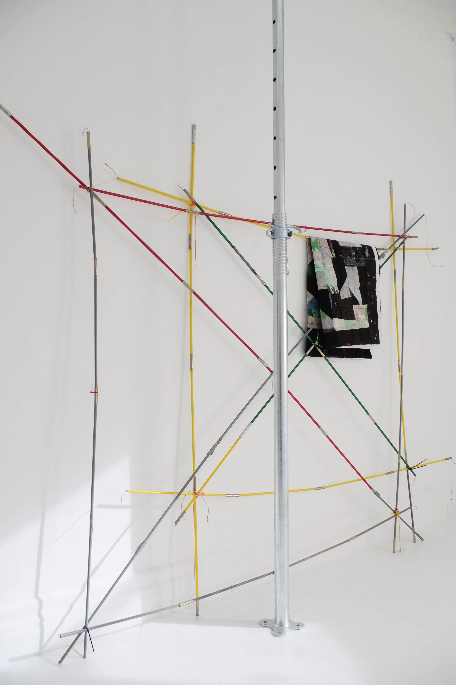 Art installation with metal rods, colorful wires, and fabric, arranged in an abstract structure attached to a metal stand against a white wall.