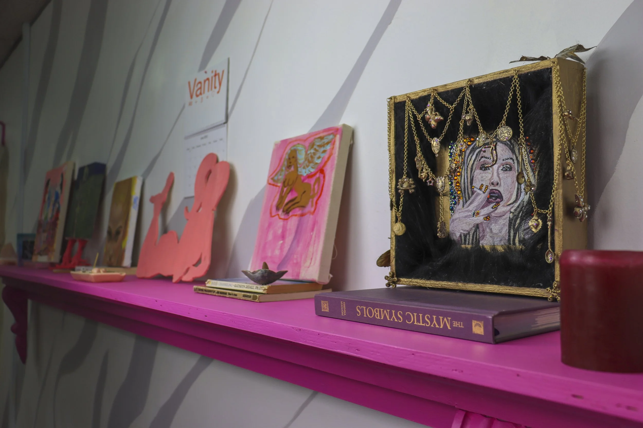 Display of artwork, jewelry, and books on a pink shelf against a wall with diagonal gray stripes. The jewelry is hanging on a framed piece featuring a portrait of a woman with painted and bejeweled details.