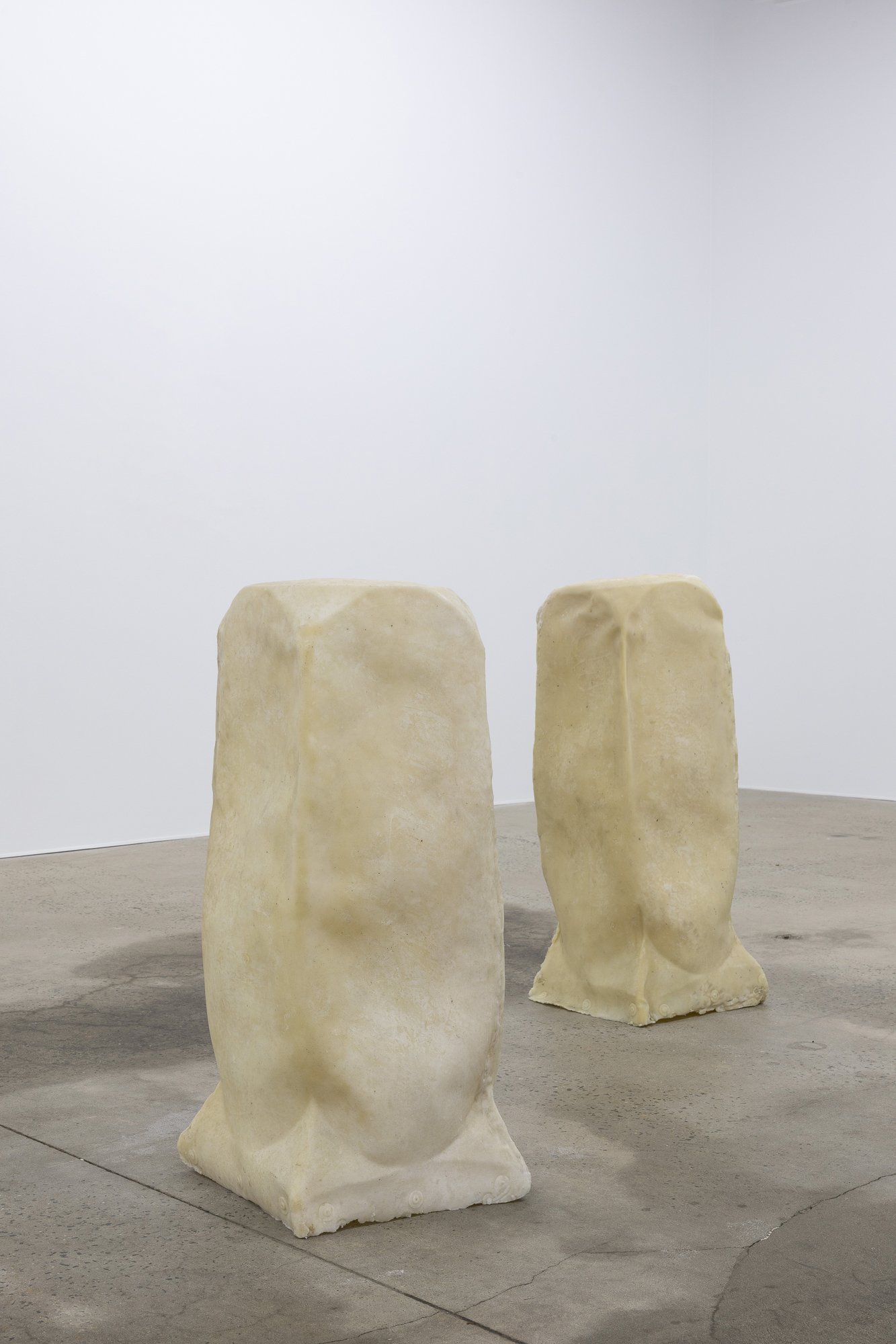 Two large, irregularly shaped, beige stone sculptures resembling rounded stools are placed on a concrete floor against a plain, white wall.