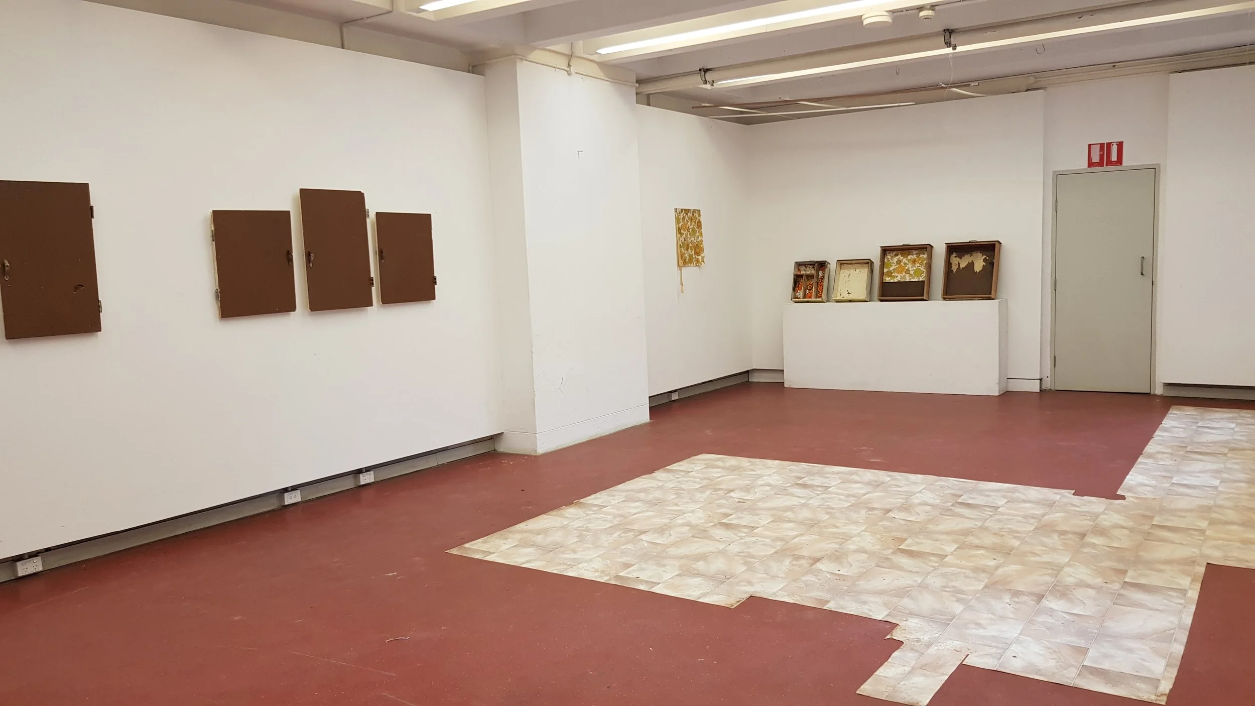 Empty art gallery with white walls, brown lockers on the left, and framed artworks on a ledge in the corner. The floor has red and beige patterned tiles, and a white door with a red fire alarm above it is visible on the right.