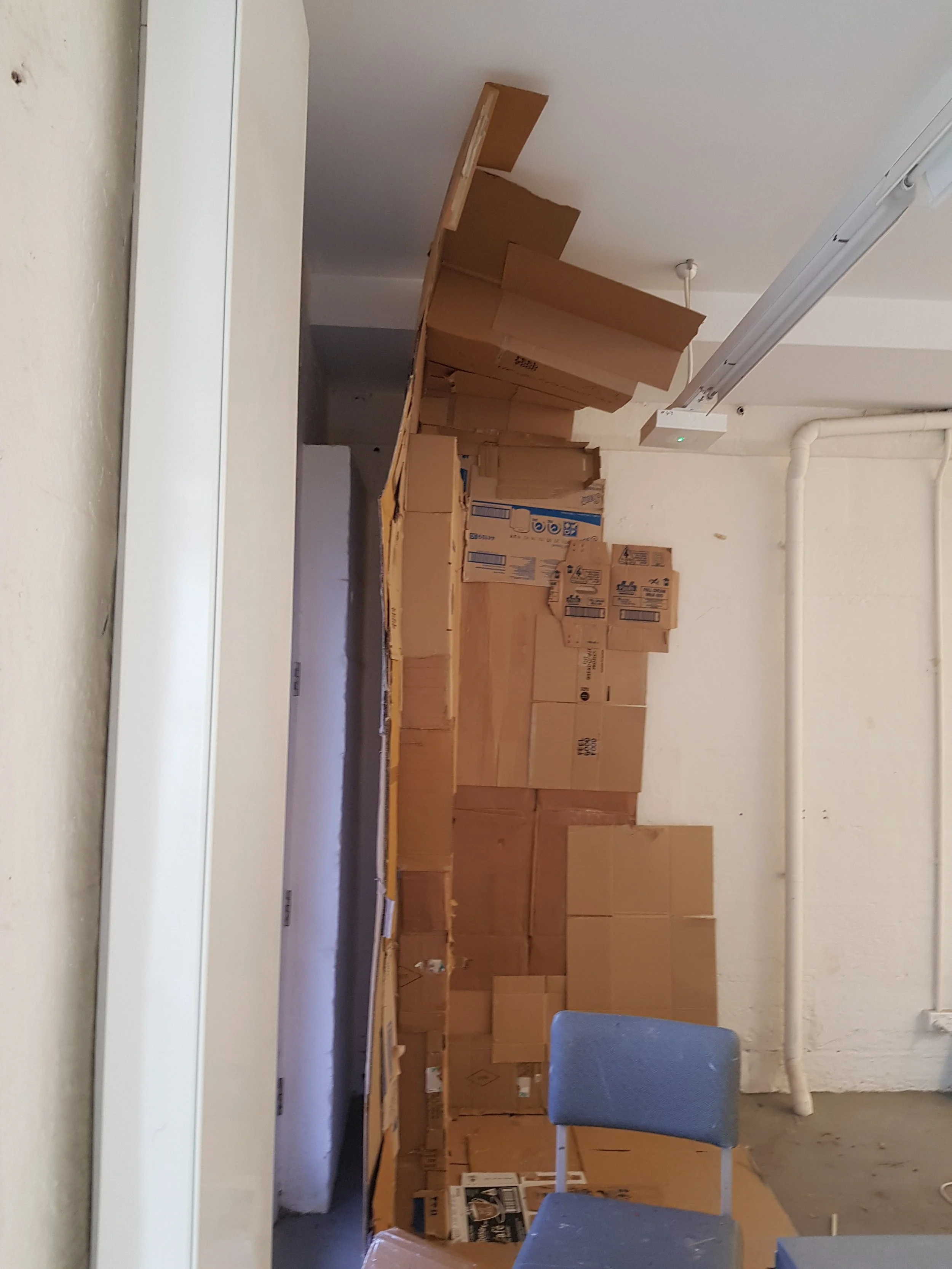 A room filled with stacked cardboard boxes, some open and others closed, with a partially assembled wooden structure and a blue chair against the wall.