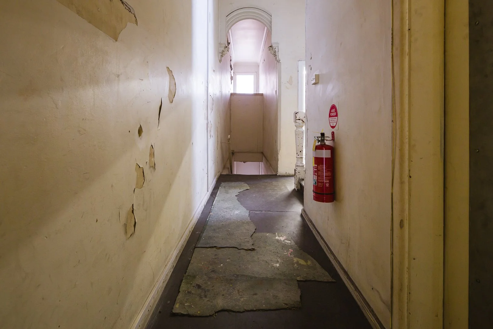 A narrow hallway with peeling yellow paint, damaged carpet remnants on the floor, and a fire extinguisher mounted on the right wall near a doorway. Light streams in from a window at the end of the hall.