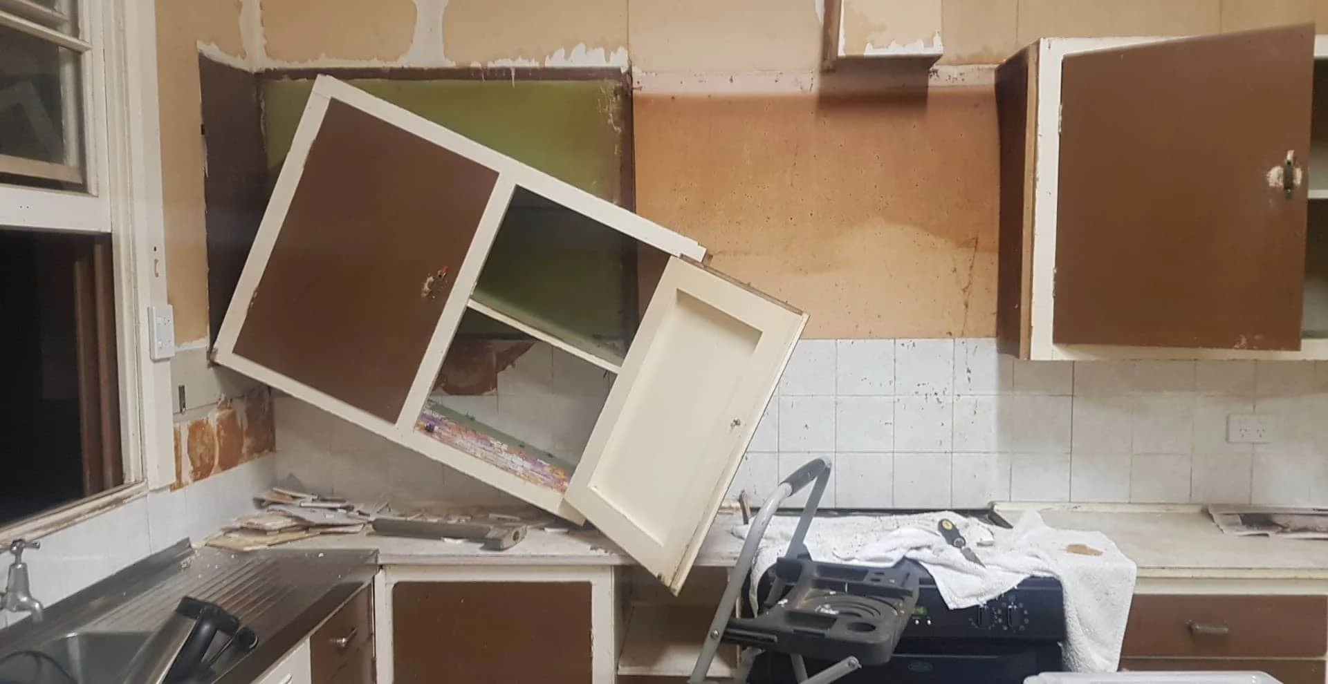 A kitchen undergoing renovation with cabinets removed, leaving exposed walls and a partially dismantled cabinet leaning against the wall. Tools and construction materials are scattered on the counter and floor, indicating ongoing work.