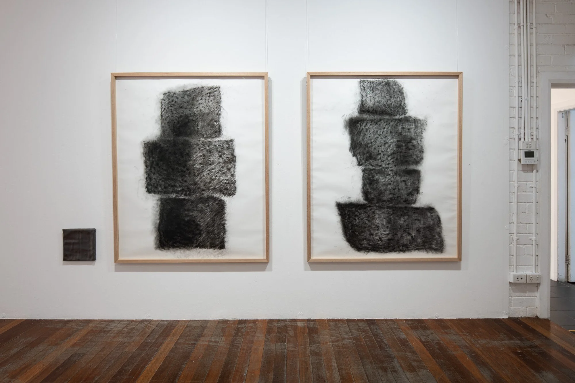 Two abstract black and gray textured art pieces in rectangular frames hanging on a white wall in an art gallery.