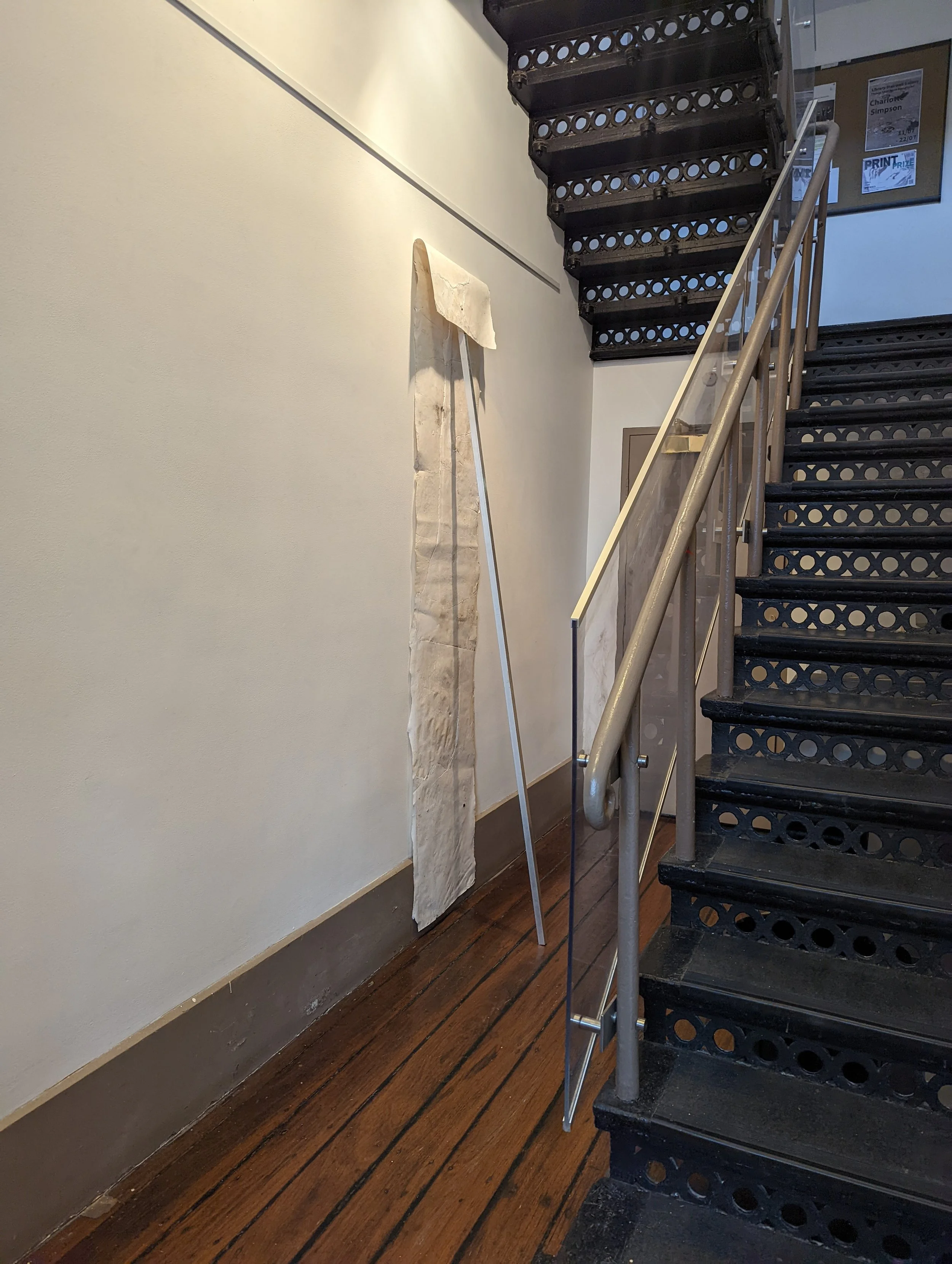 Interior view of a staircase with a white wall and a wooden floor, a piece of fabric hanging on a pole, and some posters or notices on the wall.