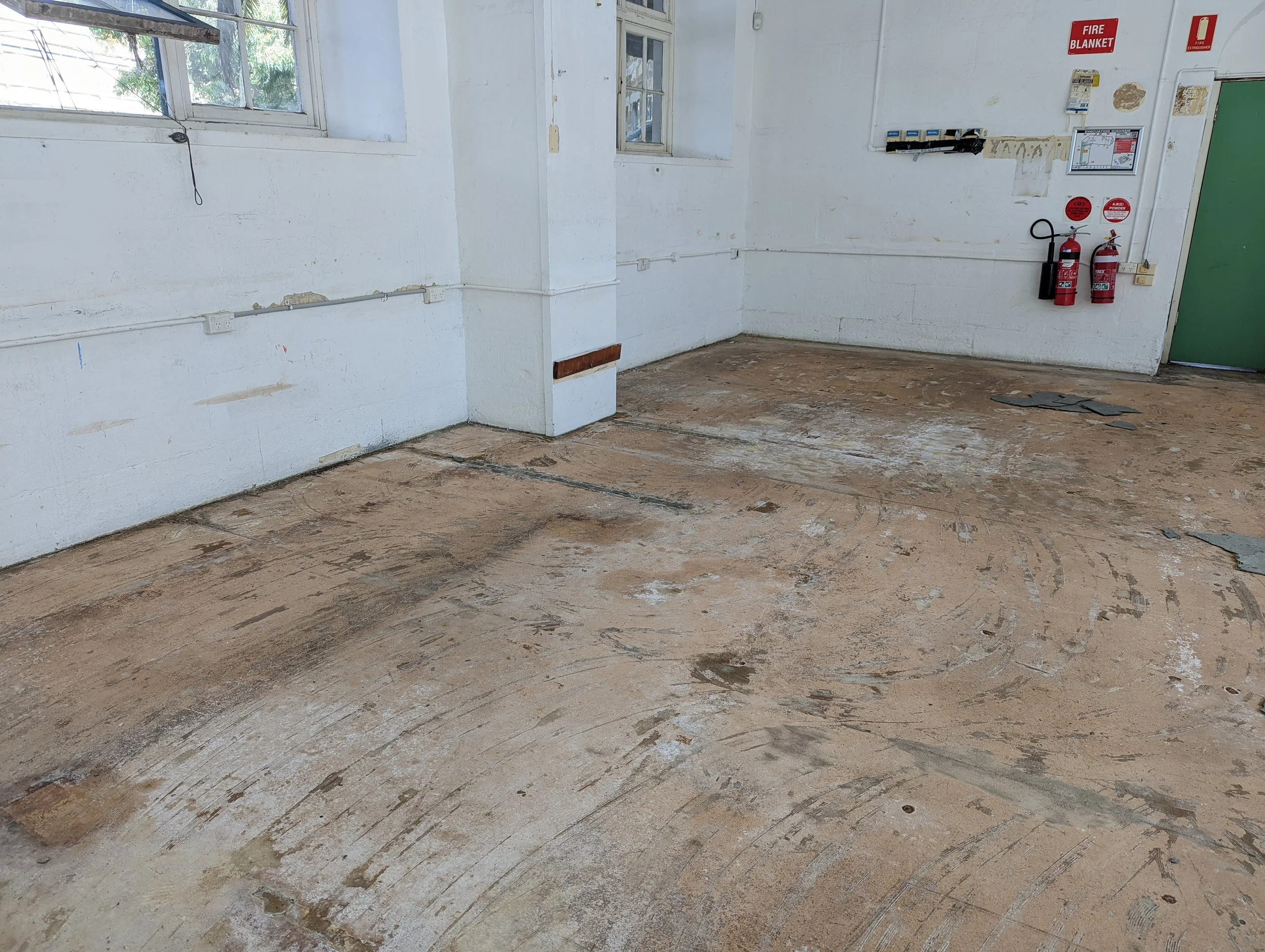 Empty room with bare wooden floor, white walls with some peeling paint, two small windows, and fire safety equipment including two fire extinguishers and fire blankets.