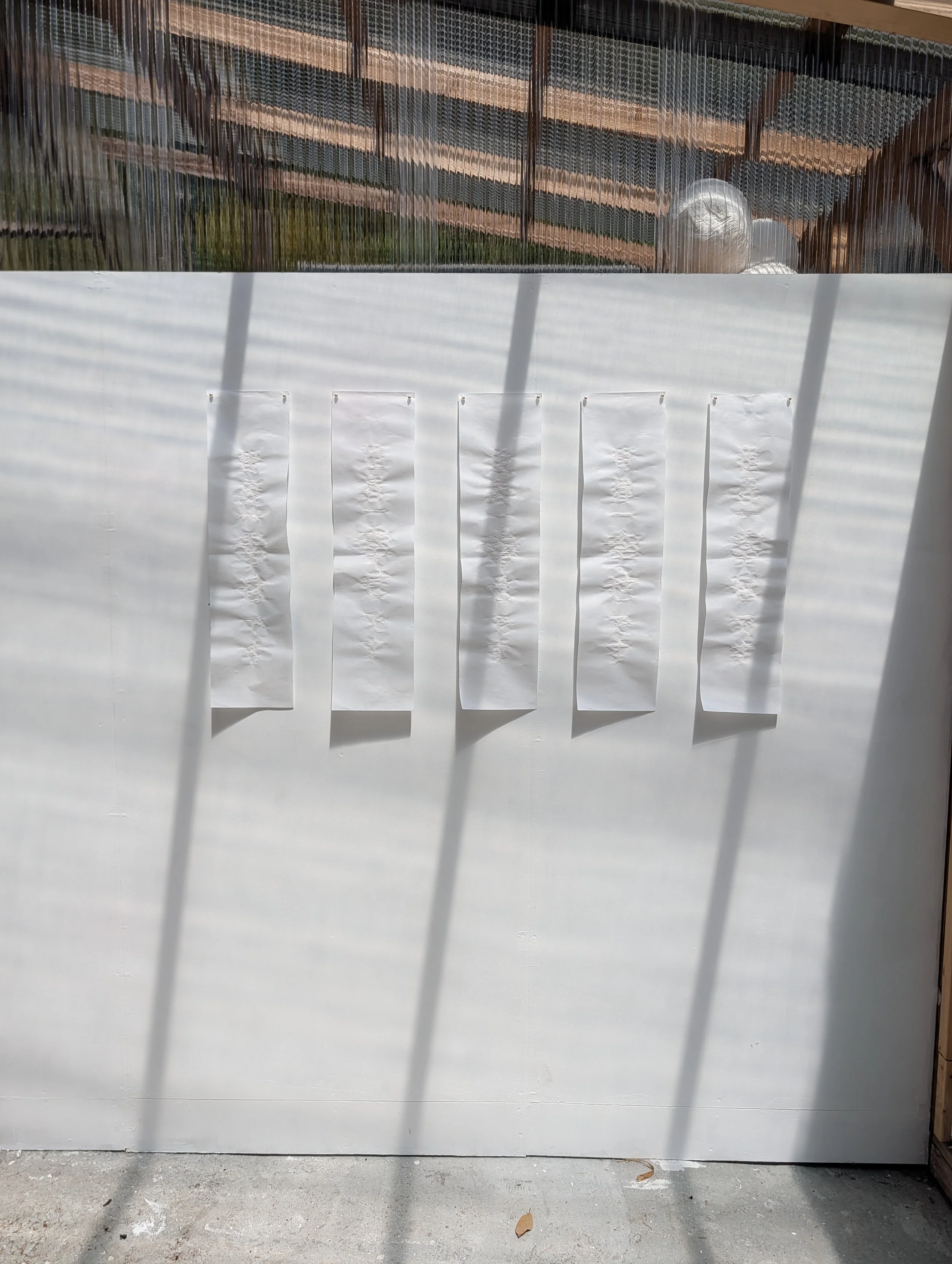 A white wall with five white paper strips taped to it, with shadows cast by overhead blinds on the wall and paper. The paper strips have some embossed or textured pattern. The top part of the image shows a reflective surface with a view of a wooden s