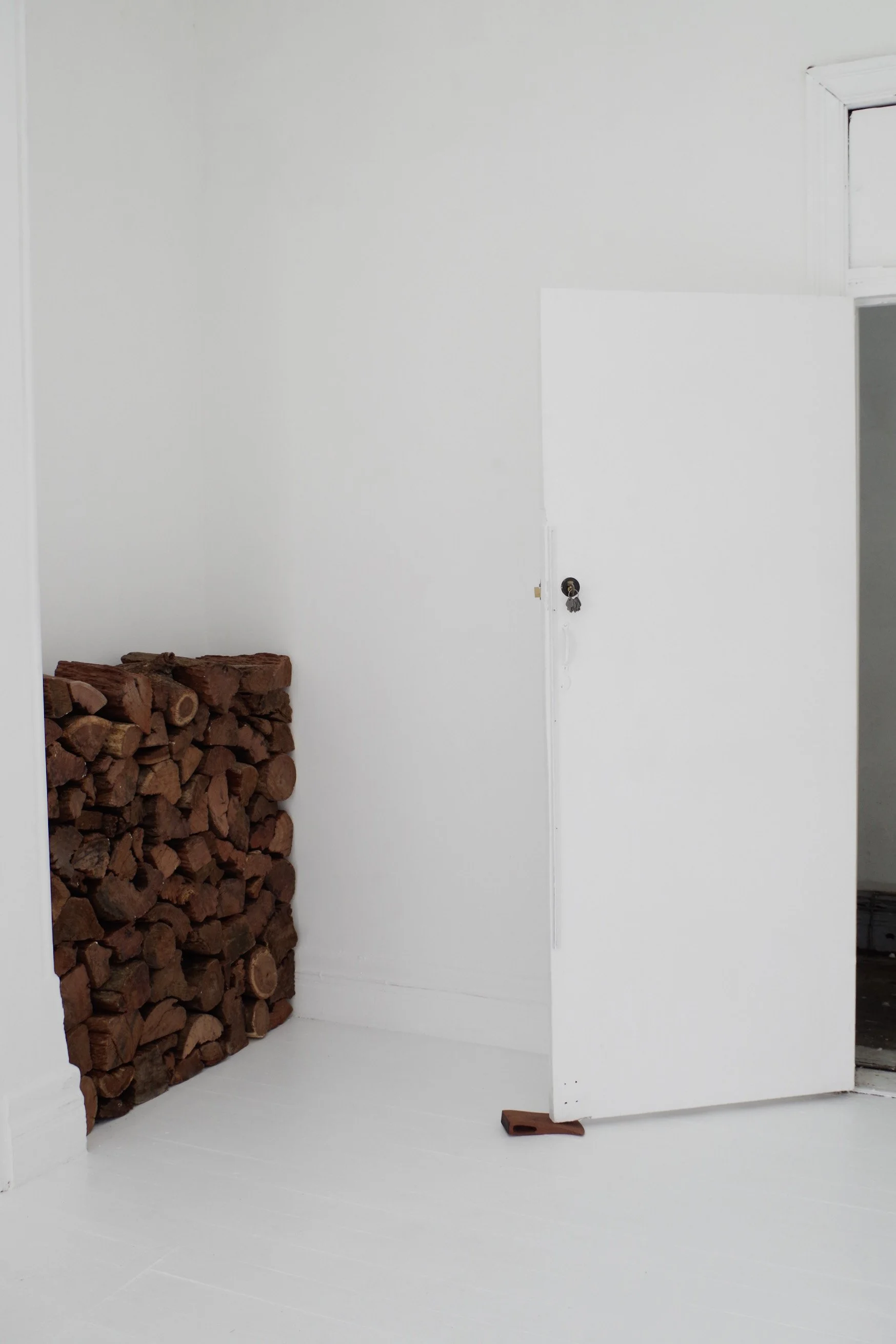 White room with a large stack of firewood and a small wooden block on the floor near the door.