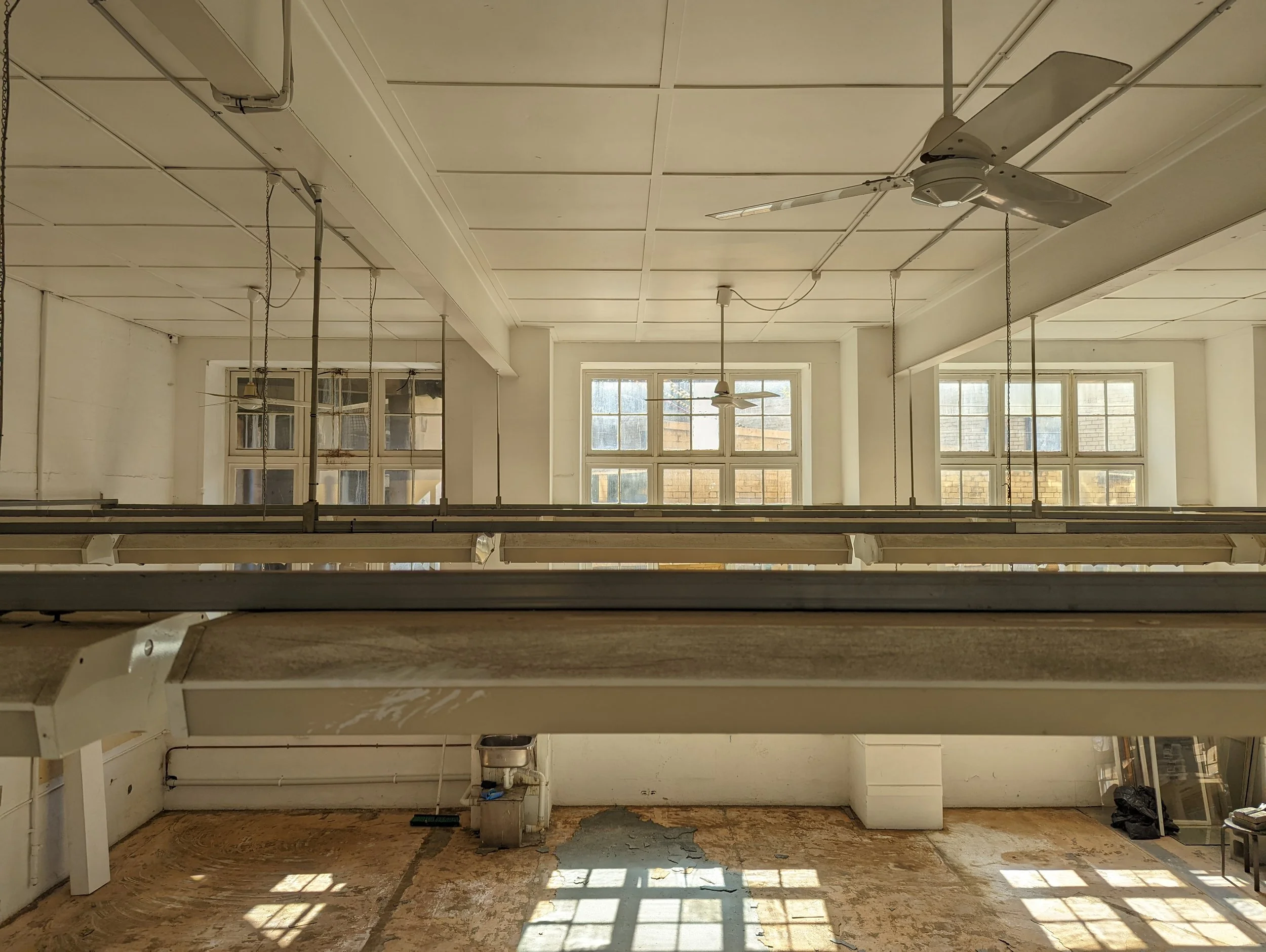 An empty room with large windows, ceiling fans, and hanging light fixtures. The floor is partially removed, exposing the underlying subfloor, indicating ongoing renovation or construction.