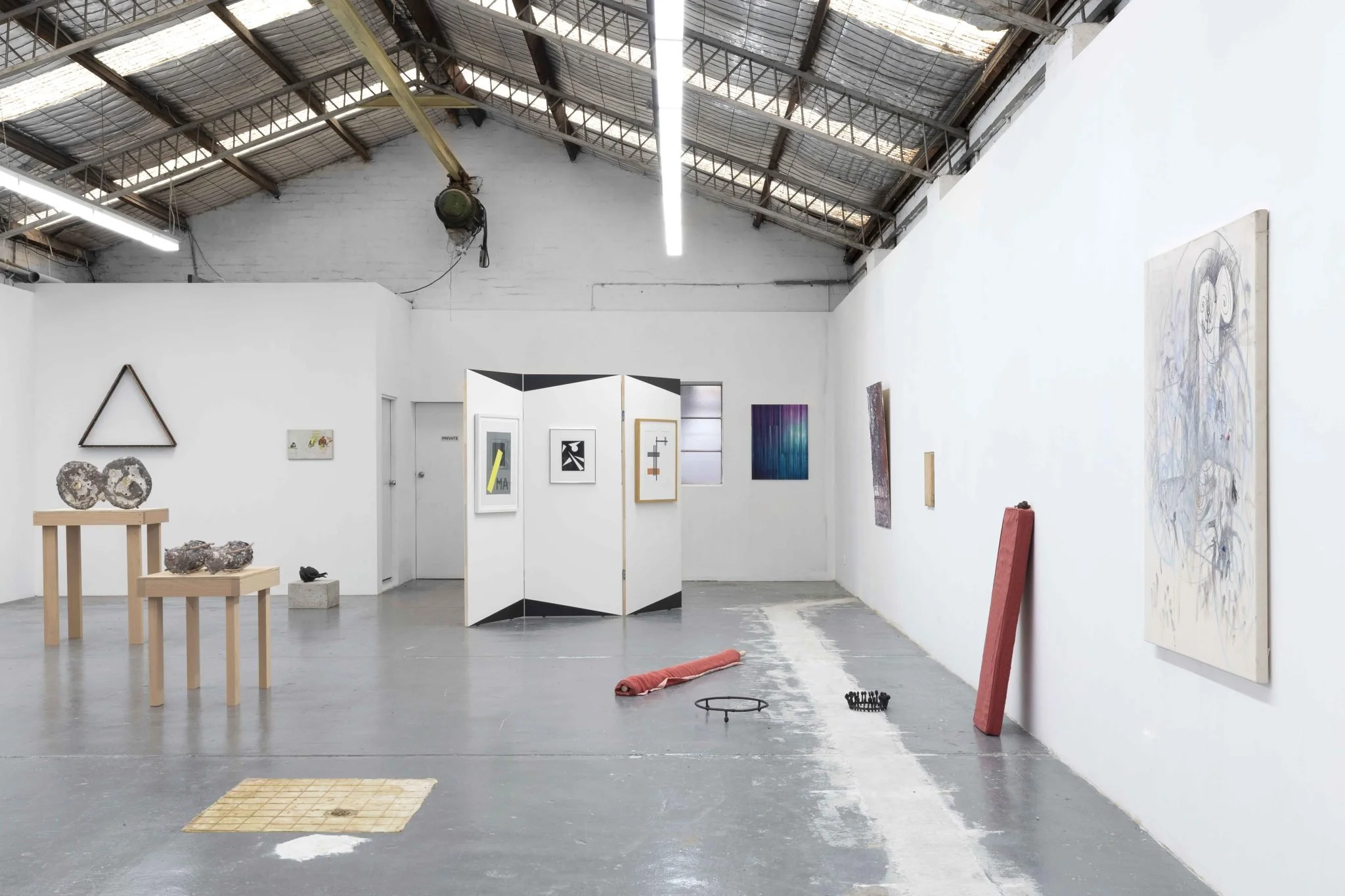 Interior of an art gallery with various modern artworks on white walls, including paintings and sculptures, and a high ceiling with exposed beams and roofing.