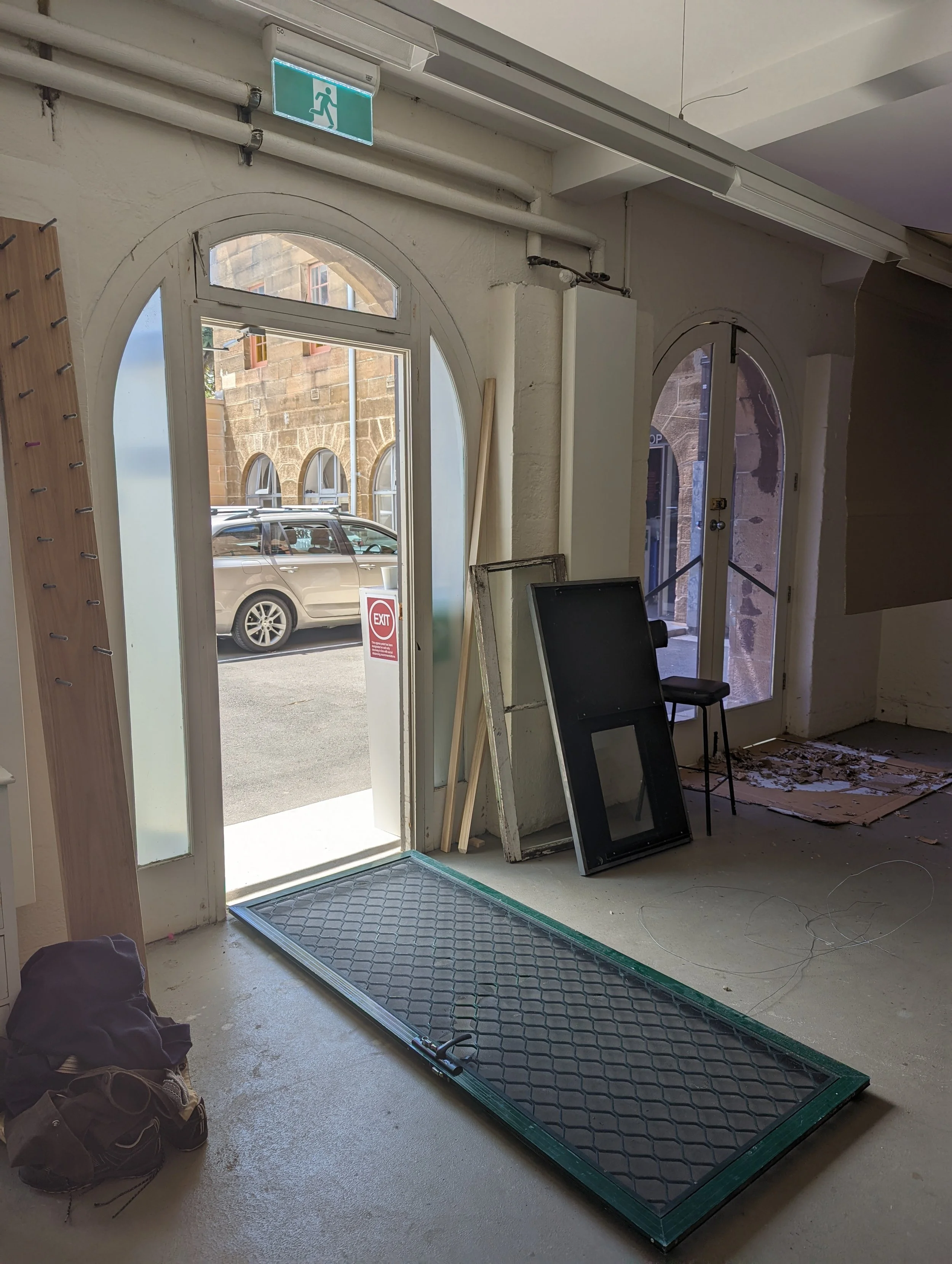 Entrance area with a glass door, a large window, and an exit sign above. A car is visible outside. The floor inside has a black metal grate, and there are some construction materials and a chair inside. The walls have some damaged areas, and there's 