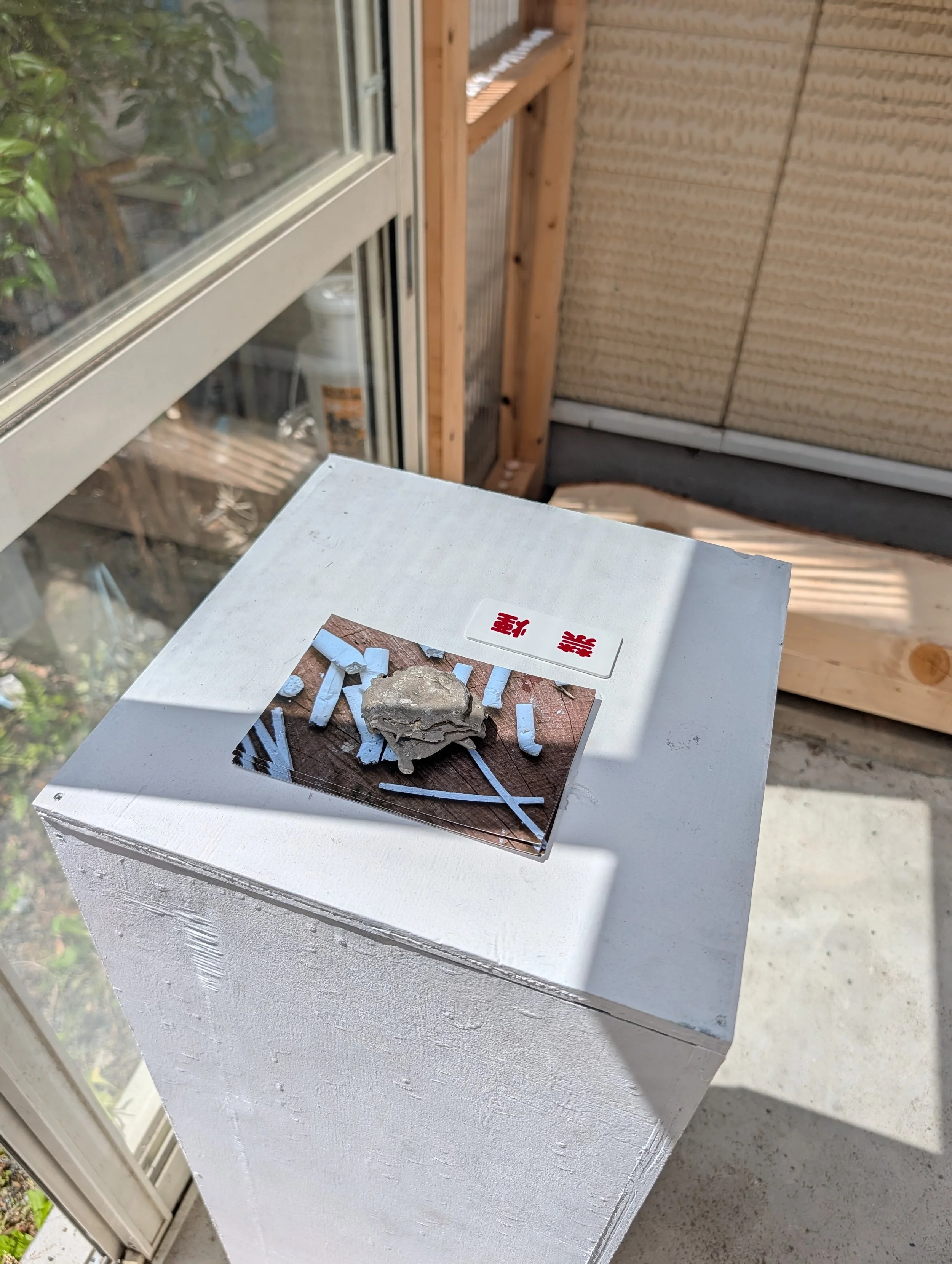 A white table or countertop has a booklet or magazine on it. The magazine cover features a rock and some blue and white building blocks. The table is beside a window, and in the background, there is a partially enclosed outdoor space with wooden fram