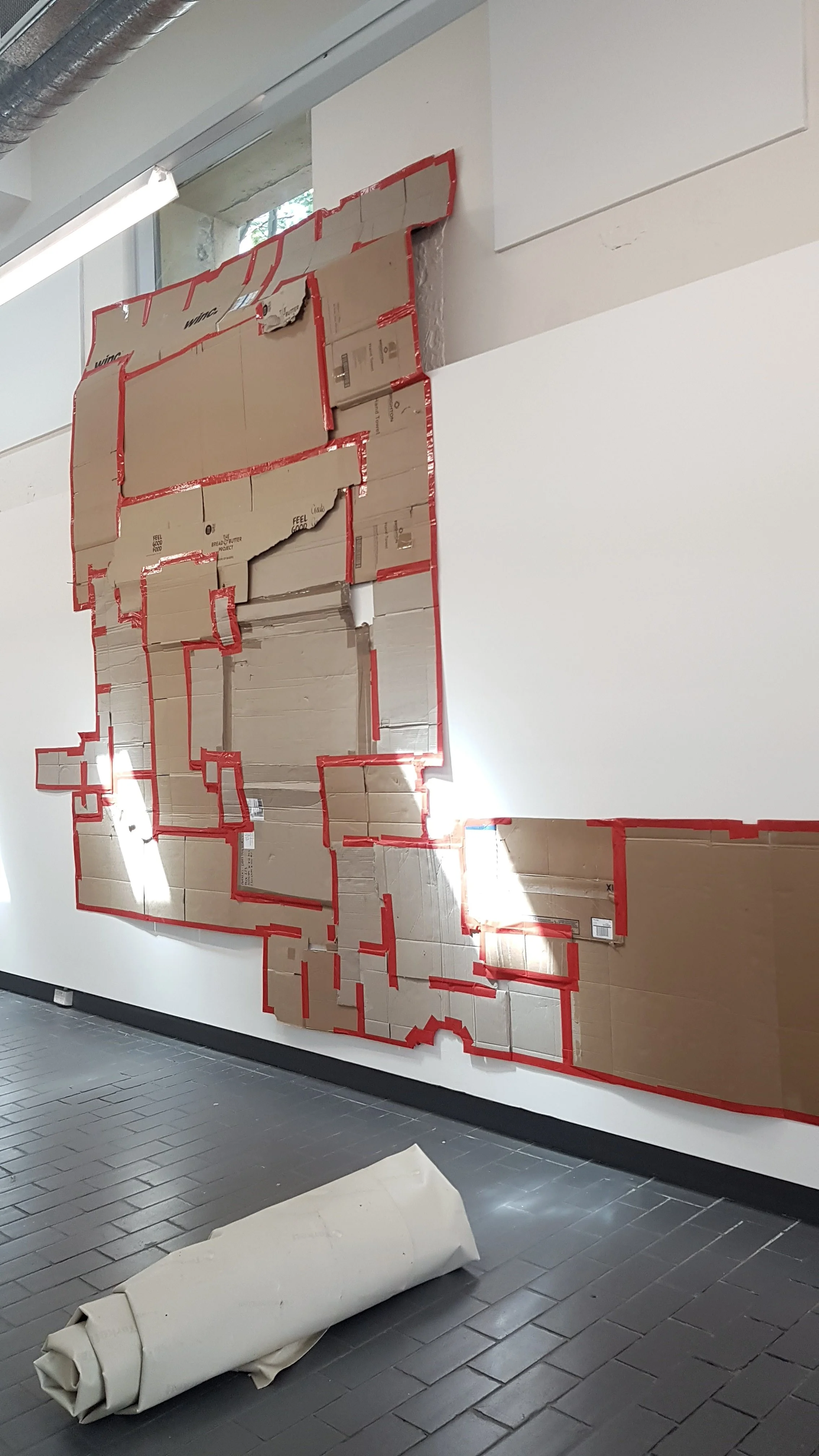 A large wall installation made of cardboard boxes arranged to form a map or design, with red tape outlining the shapes. There is a rolled-up piece of paper or cardboard on the floor in front of the wall. The setting appears to be a modern interior sp