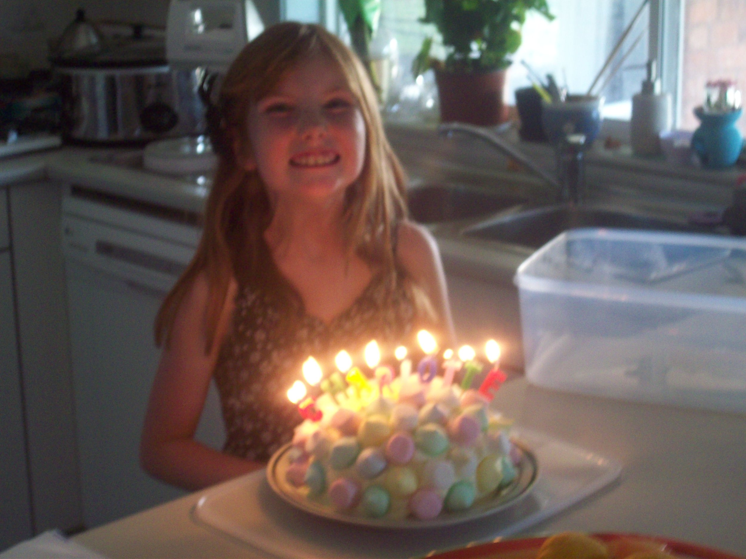 A girl smiling at a birthday celebration holding a cake with colorful candies and lit candles, standing in a kitchen near a window with plants.