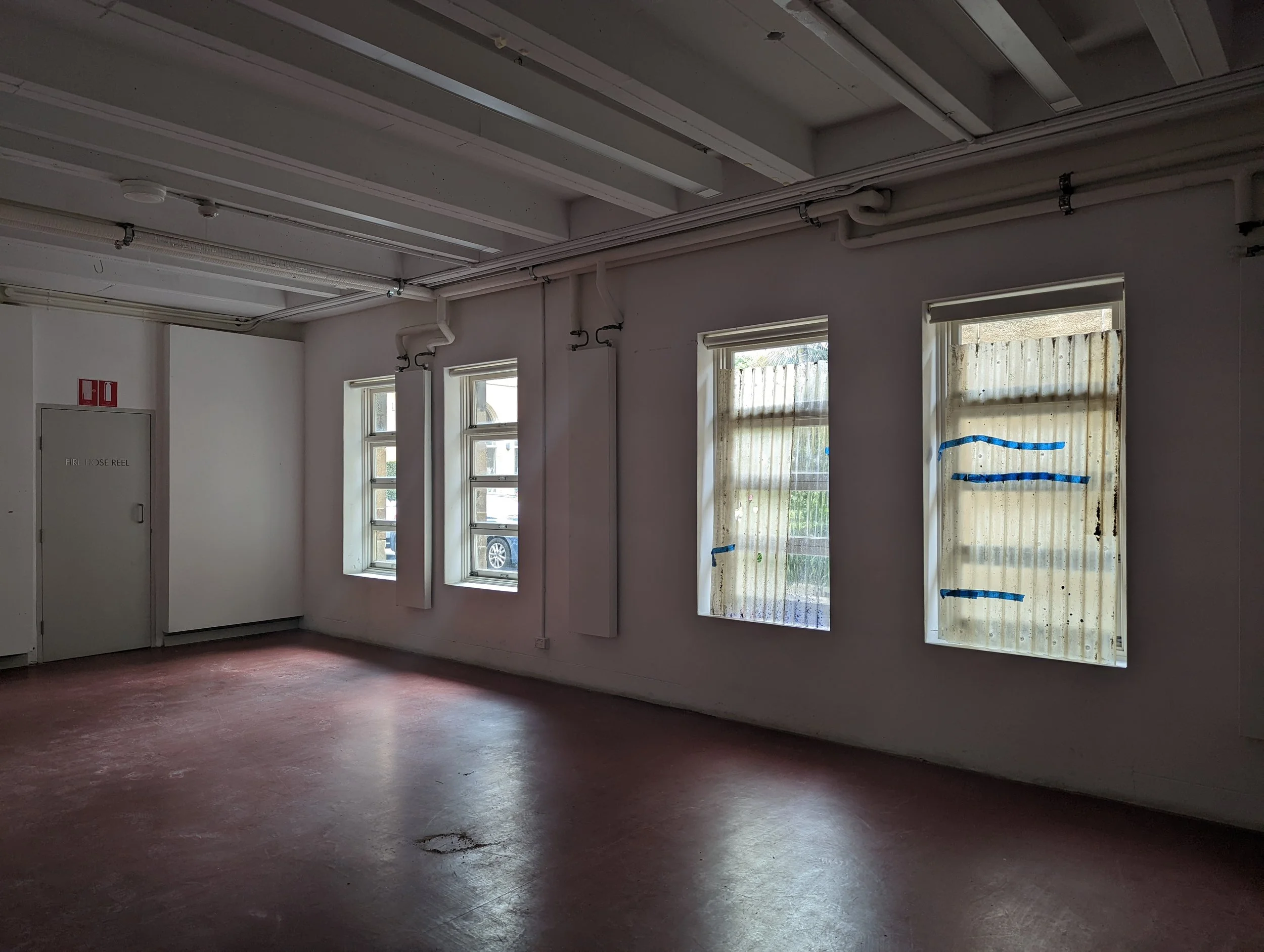 Empty room with three windows, some covered with plastic, and pipes running along the ceiling. The floor appears to be stained or worn.