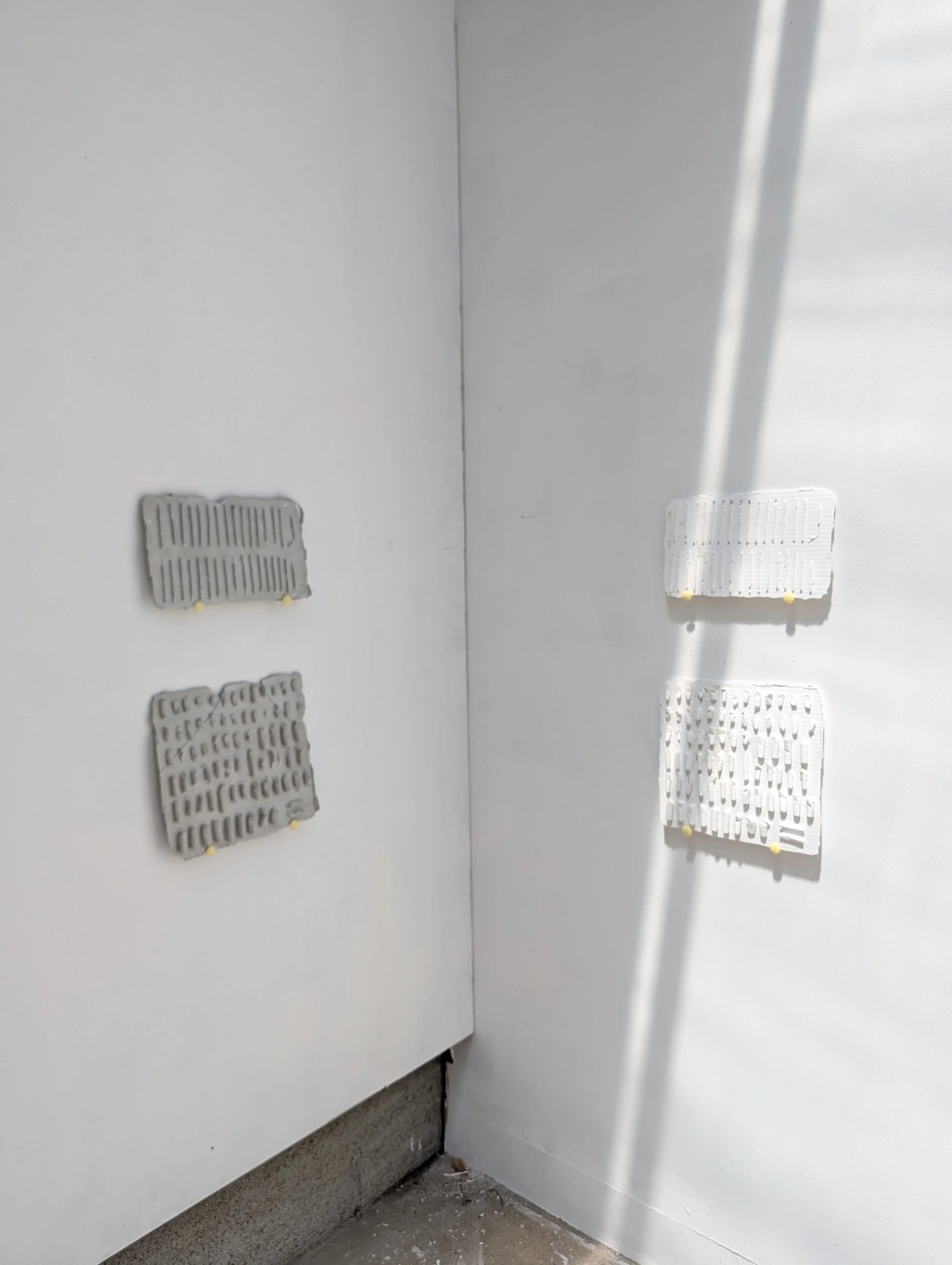 Interior corner of a room with white walls, featuring three small air vents on the lower walls and a concrete floor, with light casting shadows on the right wall.