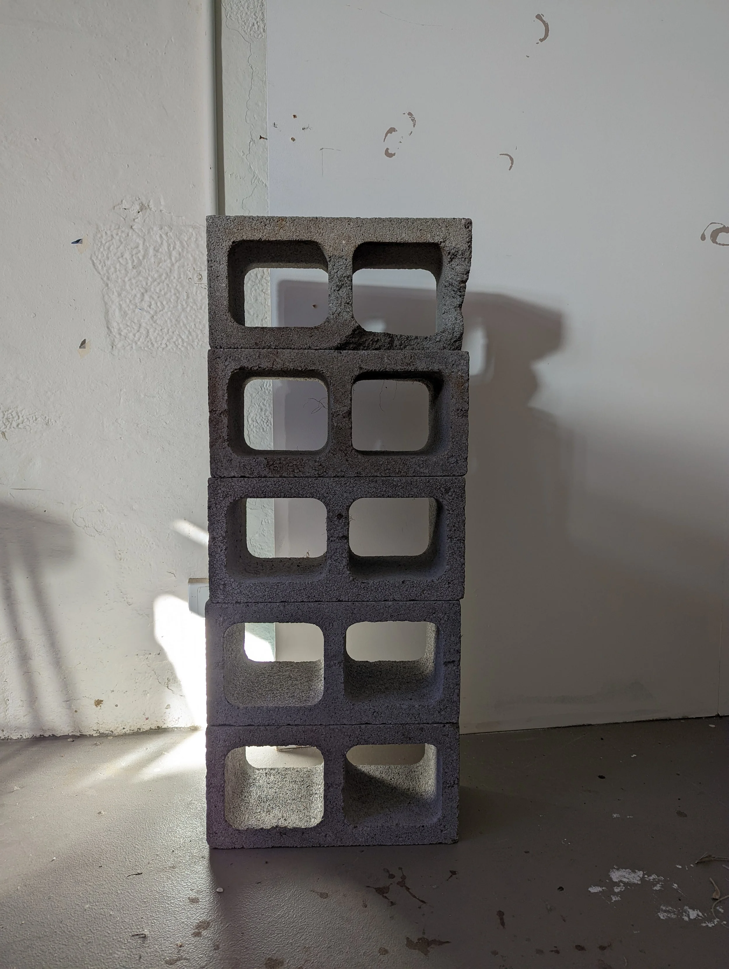 A stack of five concrete cinder blocks arranged vertically against a wall, with a window or opening visible behind the blocks.