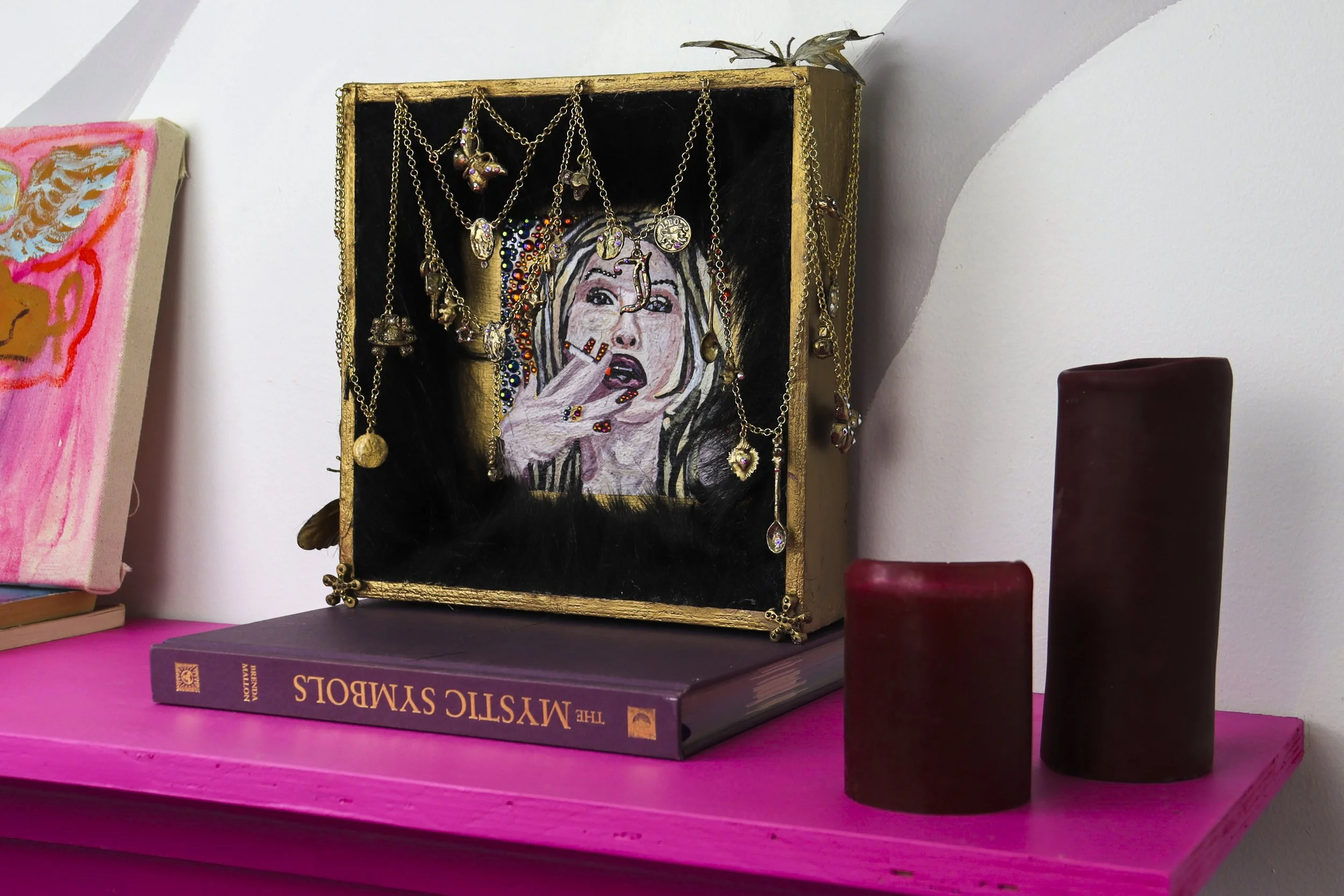 A pink shelf holding a purple book titled "The Mystic Smog" with gold lettering, topped with a jewelry box containing various necklaces with charms, a painted portrait of a woman with blonde hair making a gesture with her hand, surrounded by colorful