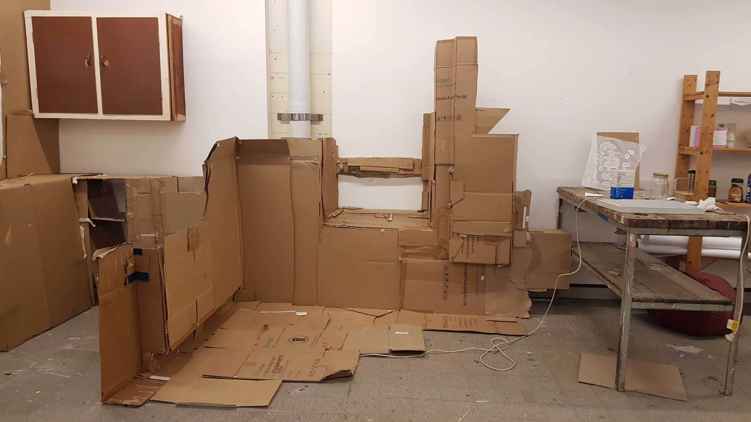 Work in progress with large cardboard structures and a work table in a room with white walls, wooden cabinets, and shelves.