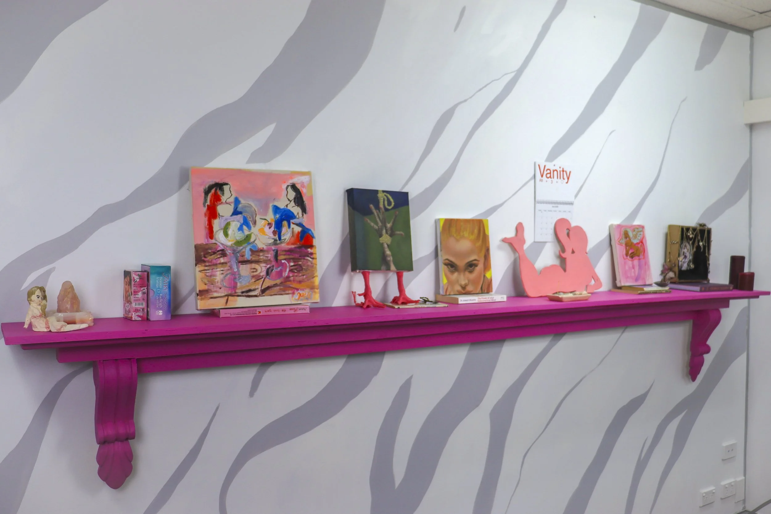 A pink shelf displaying various artwork and decorative items, mounted on a wall with gray and white abstract stripe pattern. The shelf includes paintings, a pink silhouette of a woman, a small doll, and books.