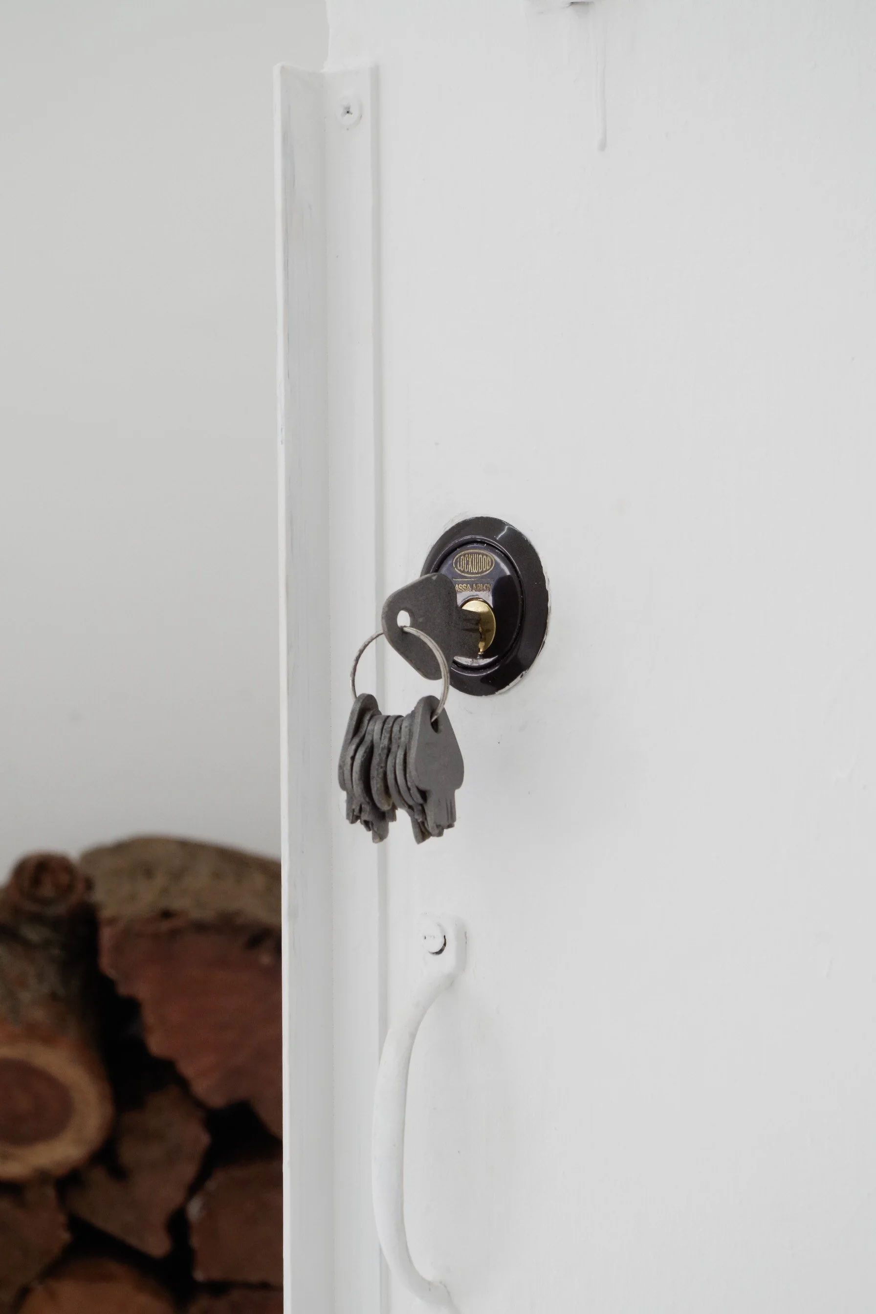 Close-up of a key inserted into a white door lock with a keychain hanging on it, background shows stacked firewood.
