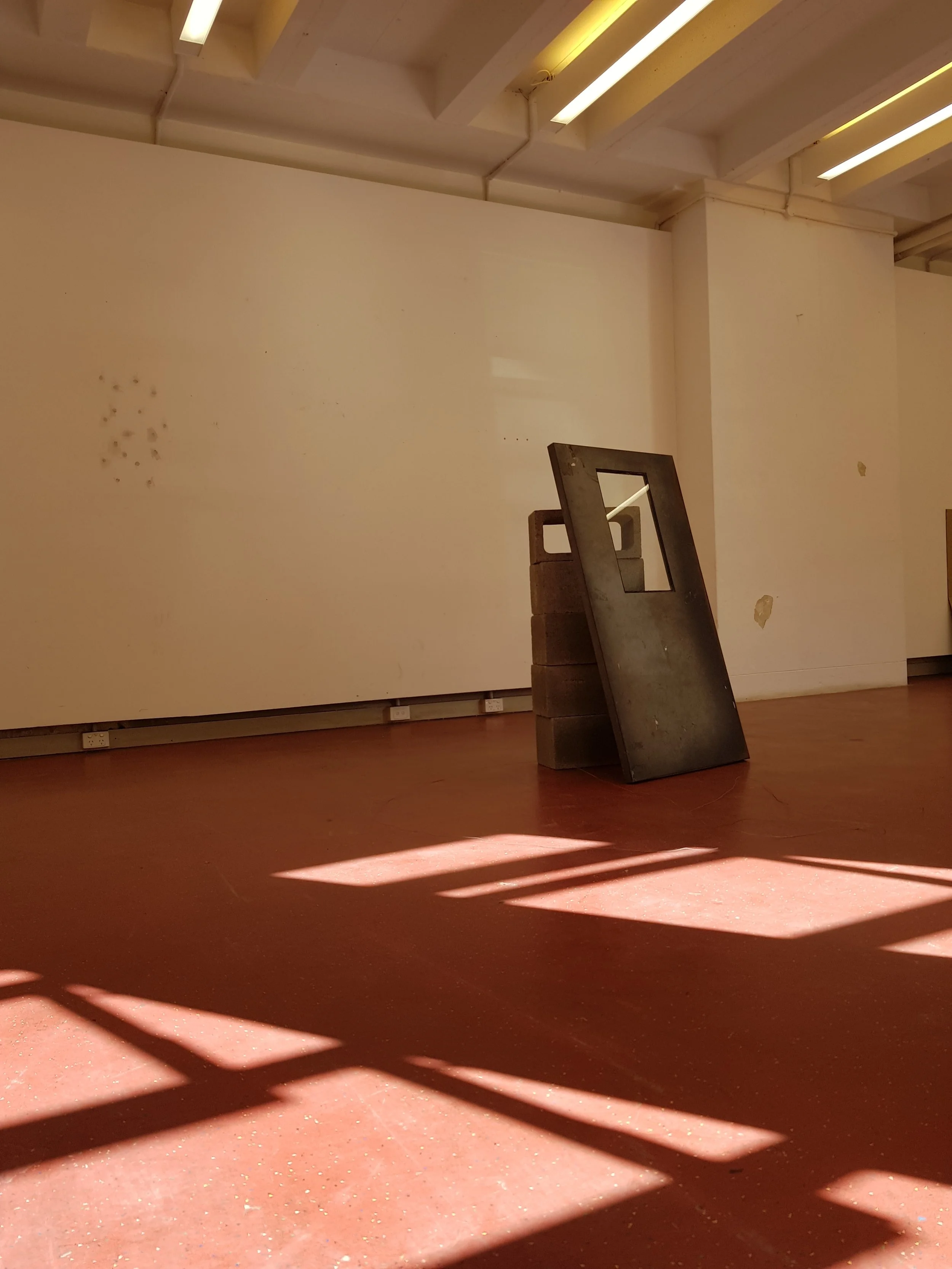 A gallery or museum interior with a white wall, a wooden floor, a shadow pattern on the floor, some small marks or holes on the wall, and a black object leaning against the wall, possibly a piece of furniture or art display.