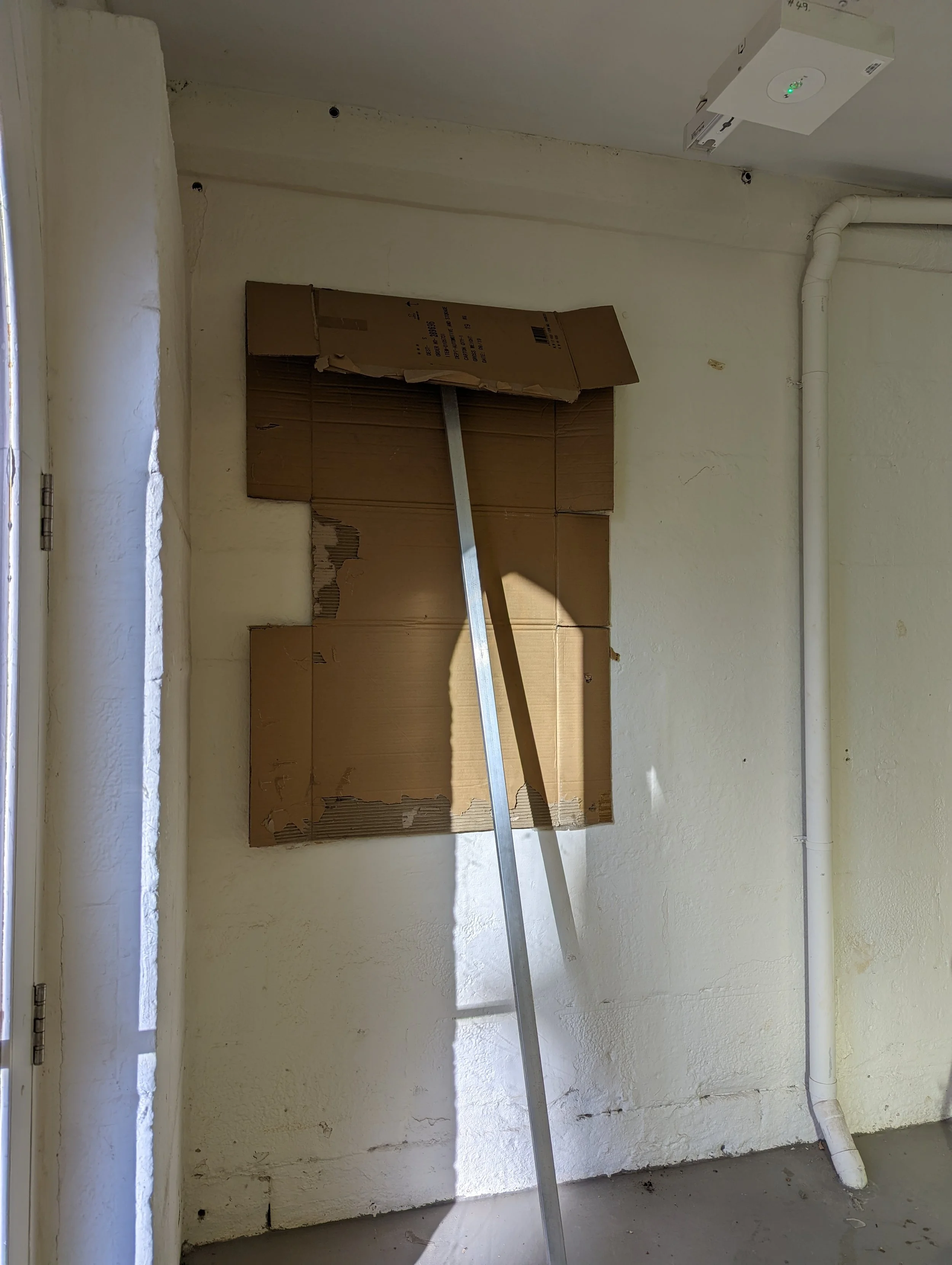 Construction or renovation site showing a corner with a drywall wall, a pipe running along the wall, a piece of cardboard covering the opening, and a metal pole leaning against the wall. There are holes in the drywall and some damage around the edges