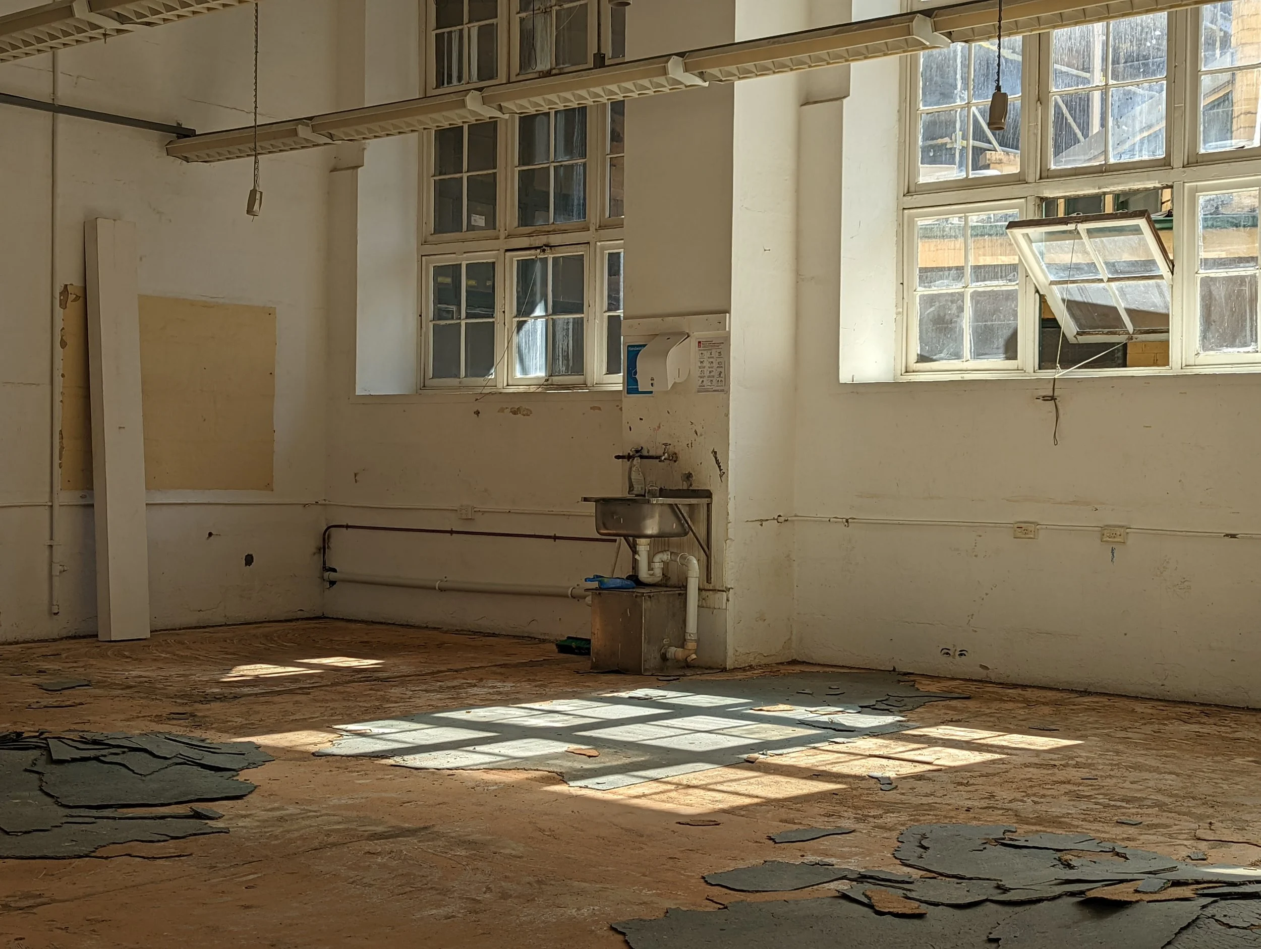 An abandoned room with large, old, multi-pane windows letting in sunlight. The floor is covered with peeling paint and debris, and the walls are unpainted and damaged. There is a small sink with exposed pipes and a paper towel dispenser on the wall.