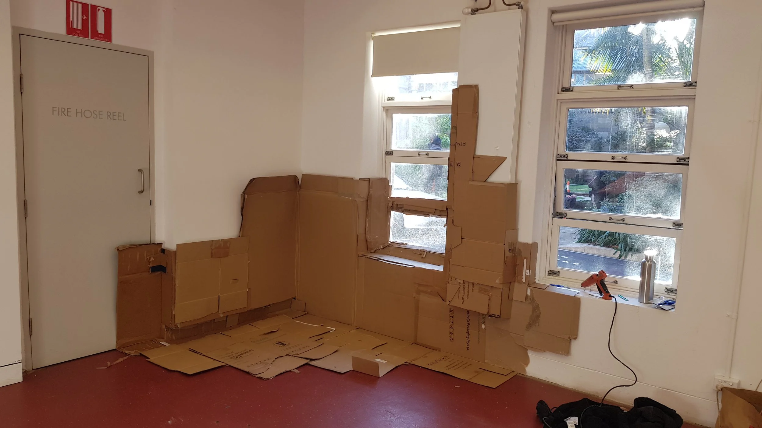 A room with three windows, some open, and brown cardboard boxes stacked along the wall. There is a red floor, a white door labeled 'FIRE HOSE REEL,' and tools like a cordless drill and a water bottle on the windowsill.