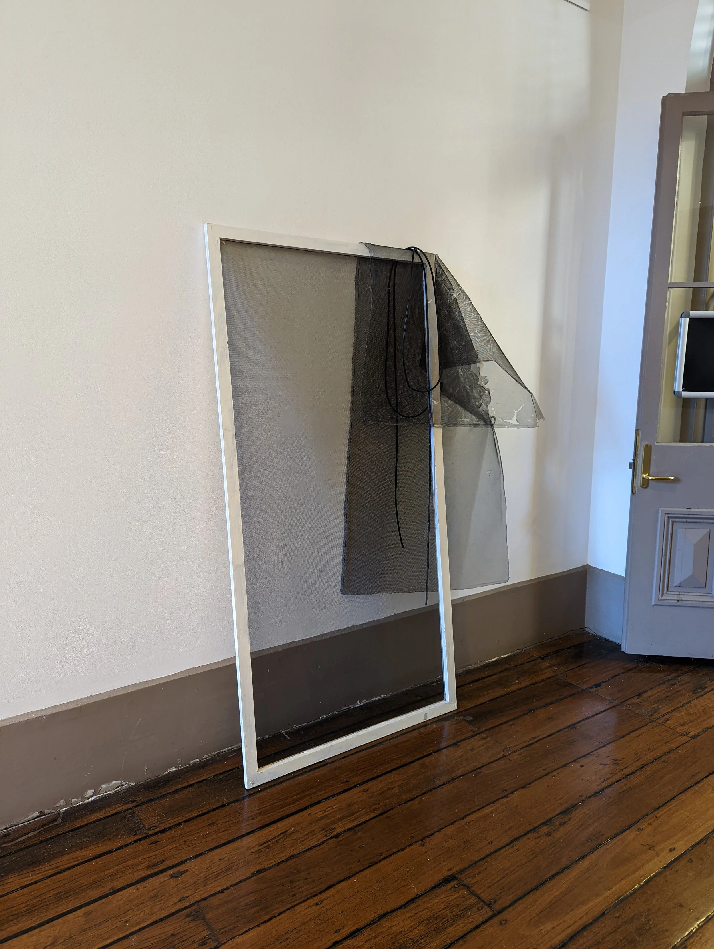 A white rectangular frame with a black mesh screen leaning against a beige wall in a room with dark wood flooring. A door with a gold handle is partially visible on the right.
