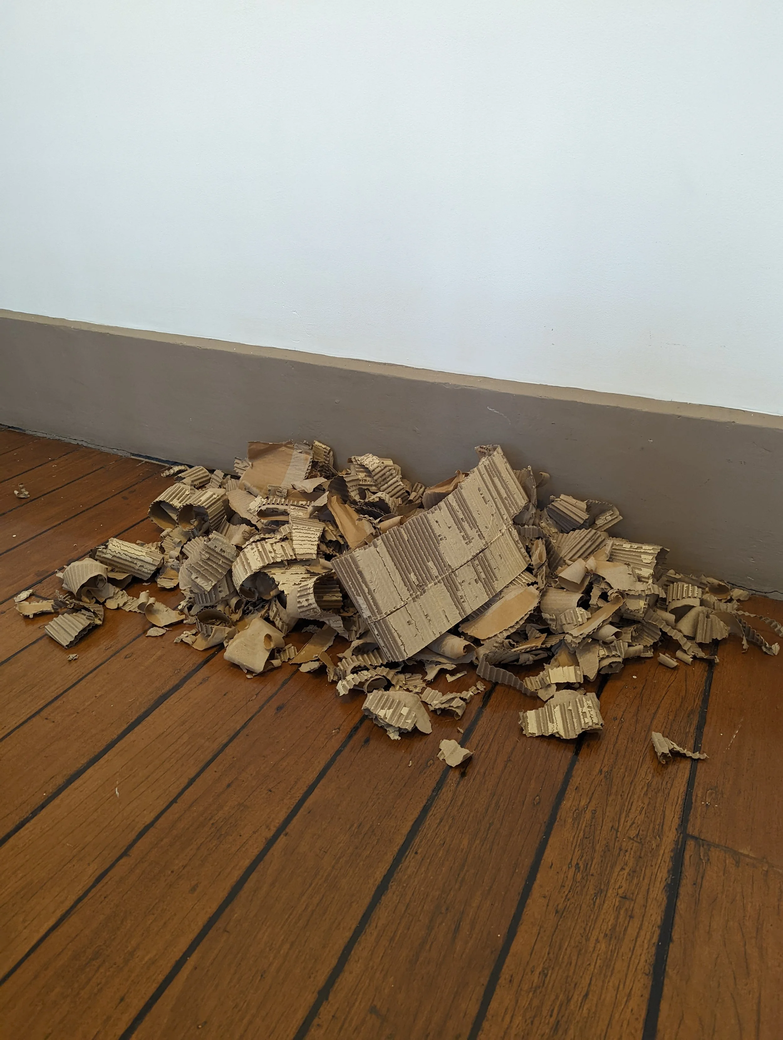 A pile of crumpled cardboard pieces on a wooden floor next to a gray baseboard and a painted white wall.