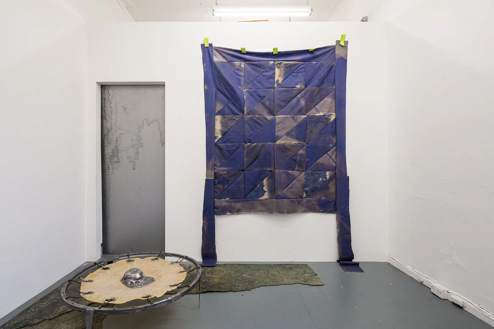 An art installation featuring a blue fabric piece with geometric patterns hung on a white wall and a metallic sculpture of a shield-like object on a circular base in the foreground.