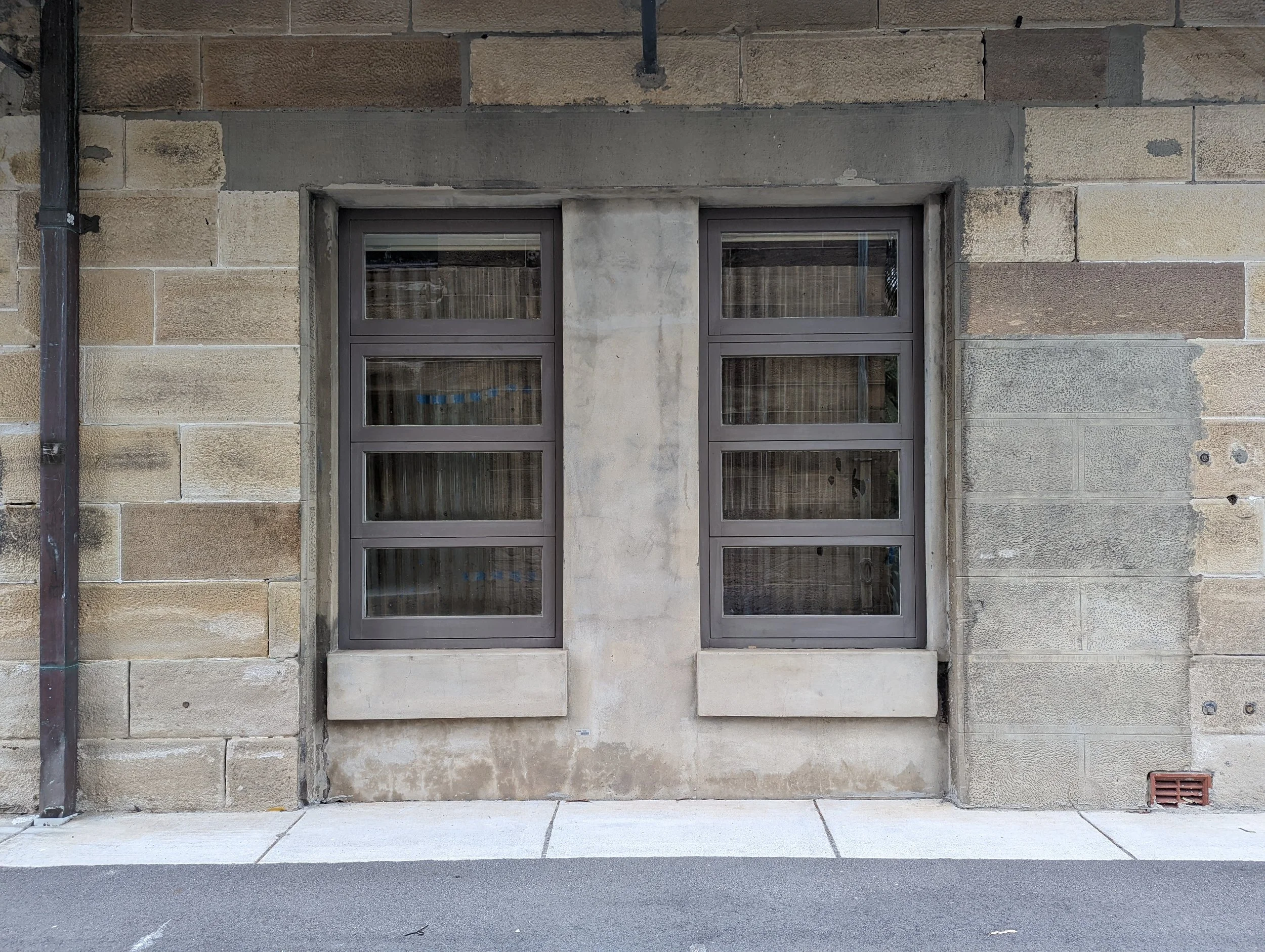 A building wall with large stone blocks and two windows with gray frames, partially covered by concrete, with a sidewalk and street in front.