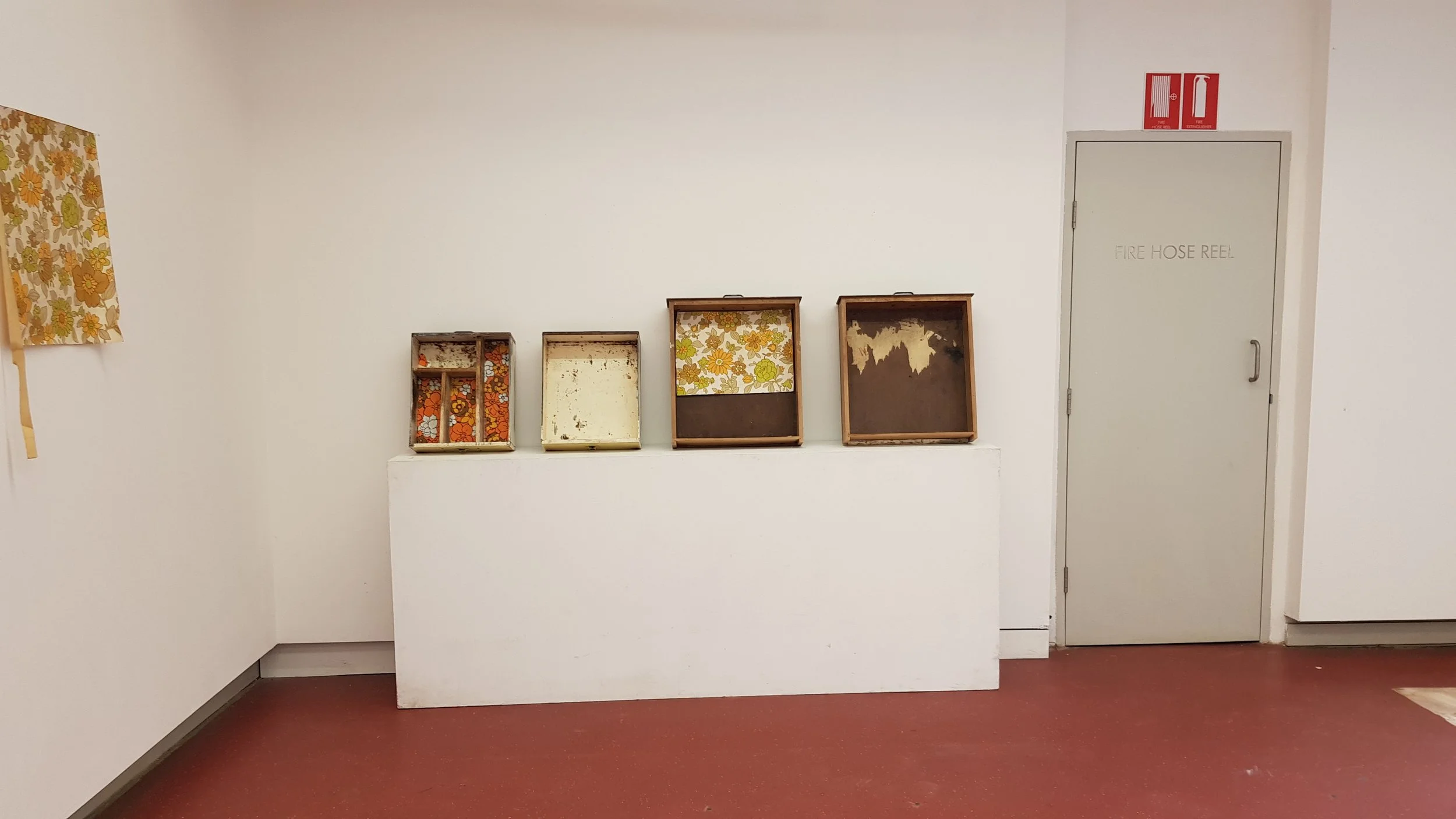 Empty vintage-style display cases with floral lining on a white wall in a room with a red floor and white walls.