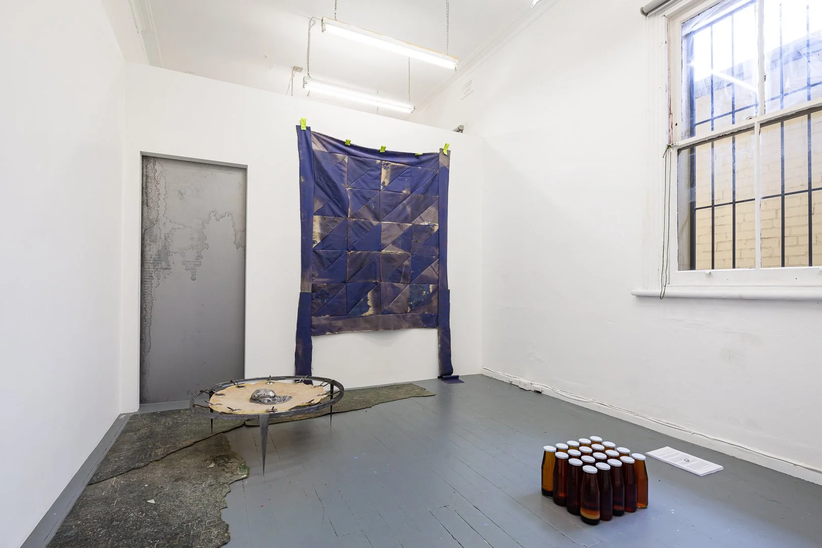 An art installation in a gallery with white walls and a gray wooden floor. It includes a large blue quilt hanging on the wall, a circular object with a reflective surface and metal frame on the floor, a cluster of brown glass bottles arranged in a ci