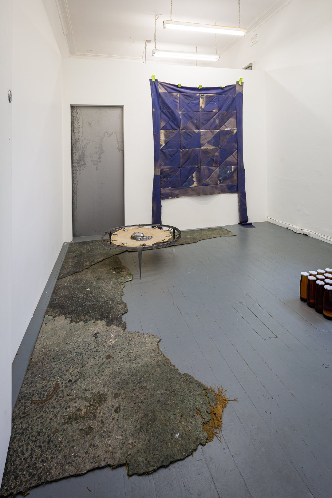 An art installation with a worn, torn rug on the floor, a large quilt hanging on the wall, a suspended circular sculpture with a small animal skull in the center, and a group of amber glass bottles on the floor in a gallery with white walls and gray 
