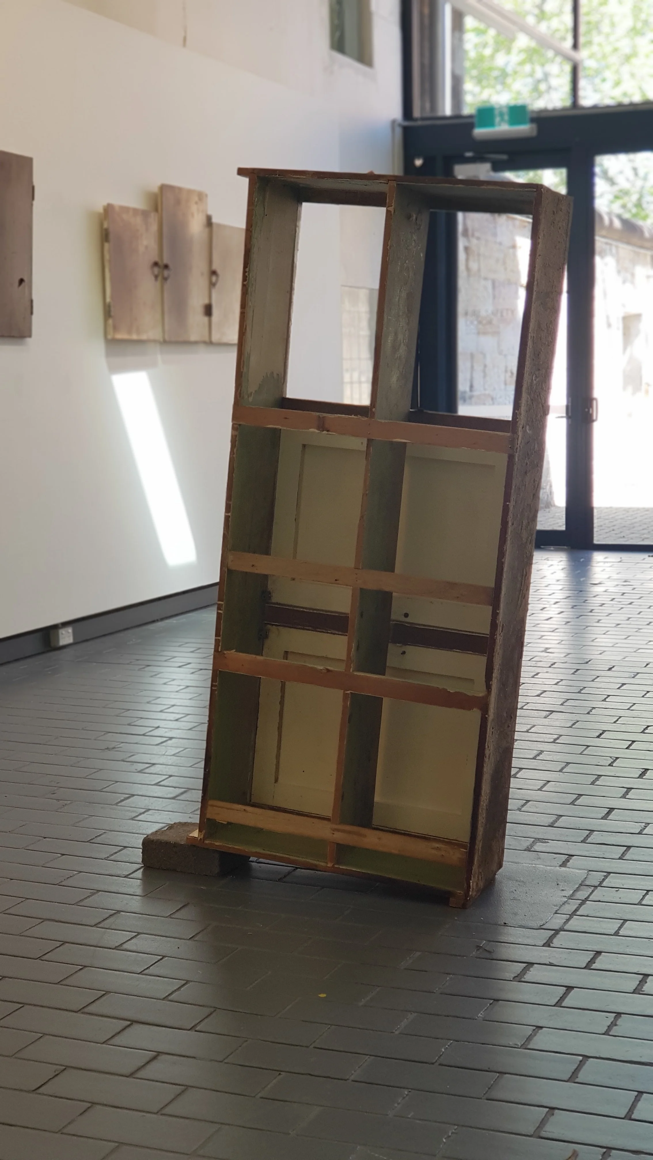 An empty wooden display shelf standing on a tiled floor in front of a glass door, with natural light coming through the door.