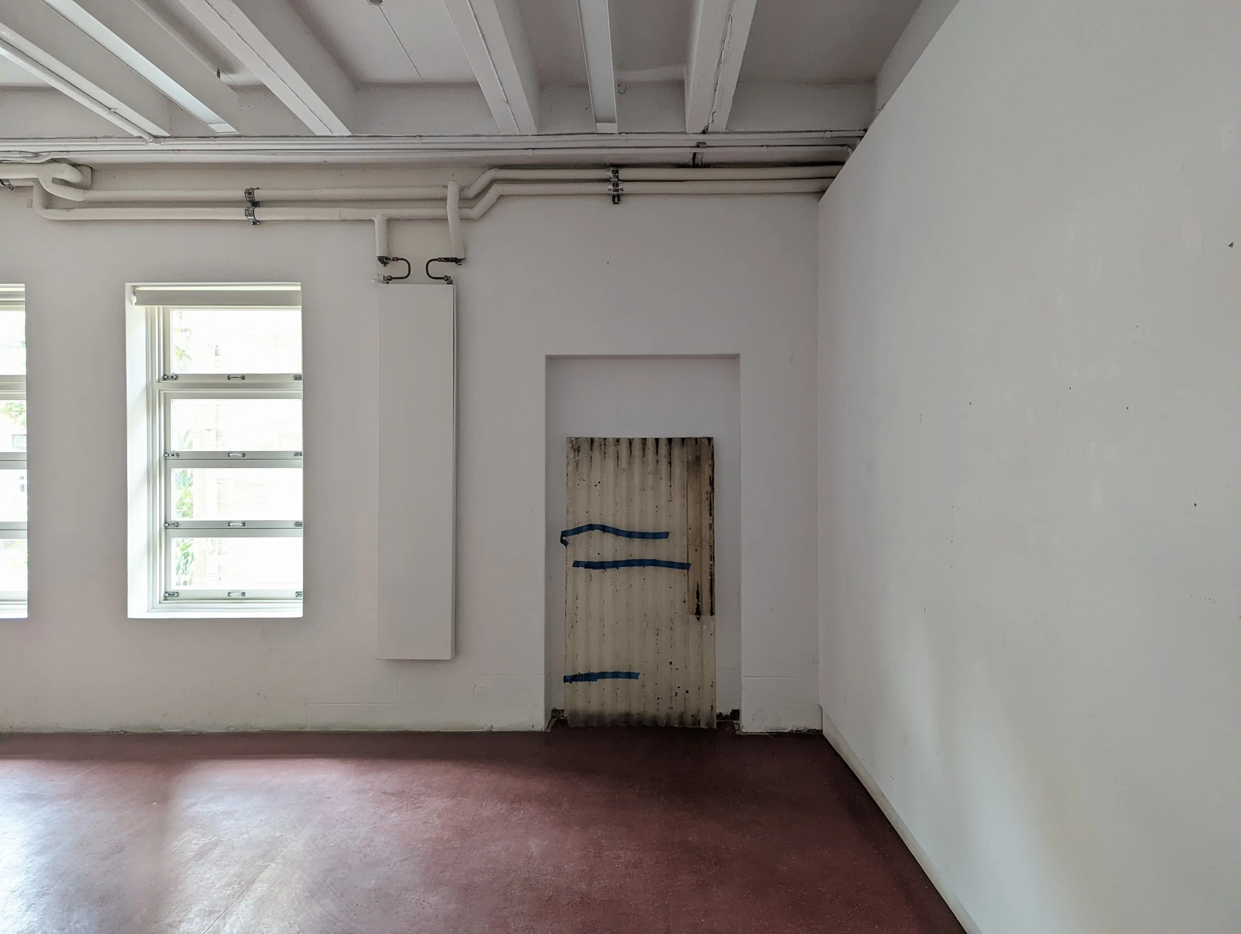 Empty room with three double-hung windows, white walls, and a section with wiring or pipes partially covered by blue painter's tape.