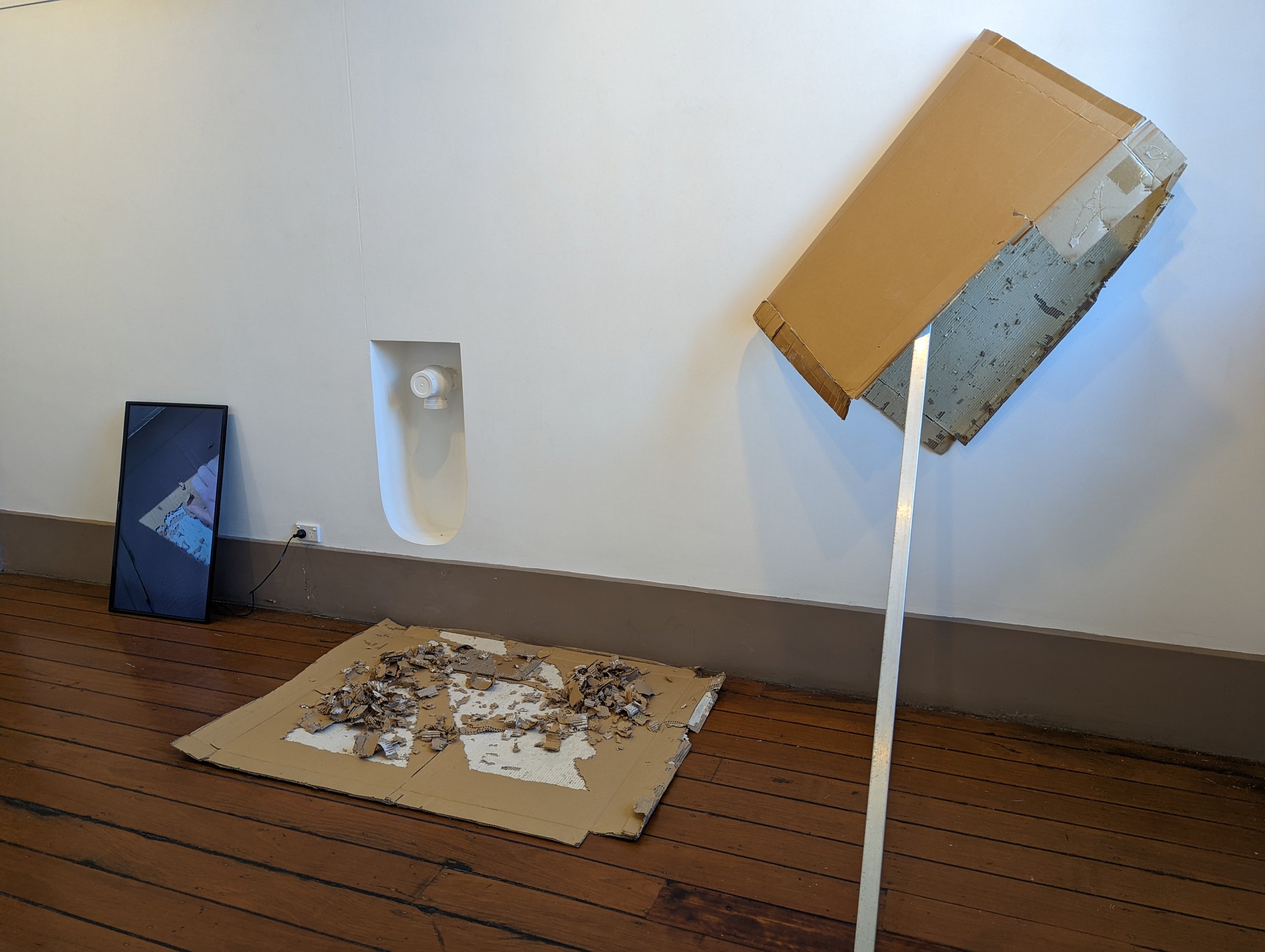 A damaged cardboard box tipped over on a stand, with torn pieces on a flattened cardboard underneath. A mirror and a wall with a smoke detector are visible in the room.