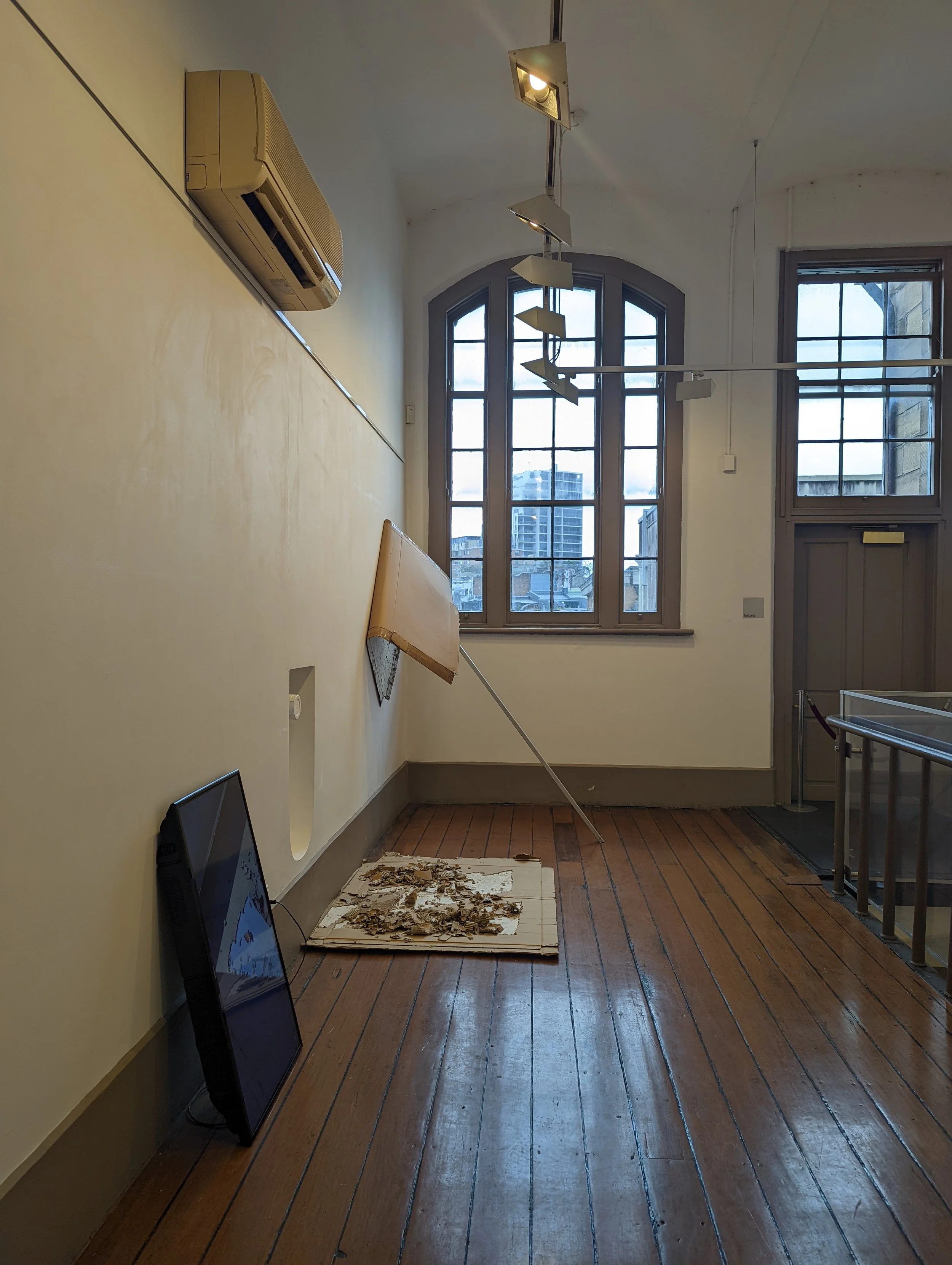 Interior of a room with tall windows, wooden floor, and a yellow wall. There are two black monitors leaning against the wall, a broken tile with debris on the floor, a wall-mounted air conditioning unit, and a hanging light fixture with multiple bulb