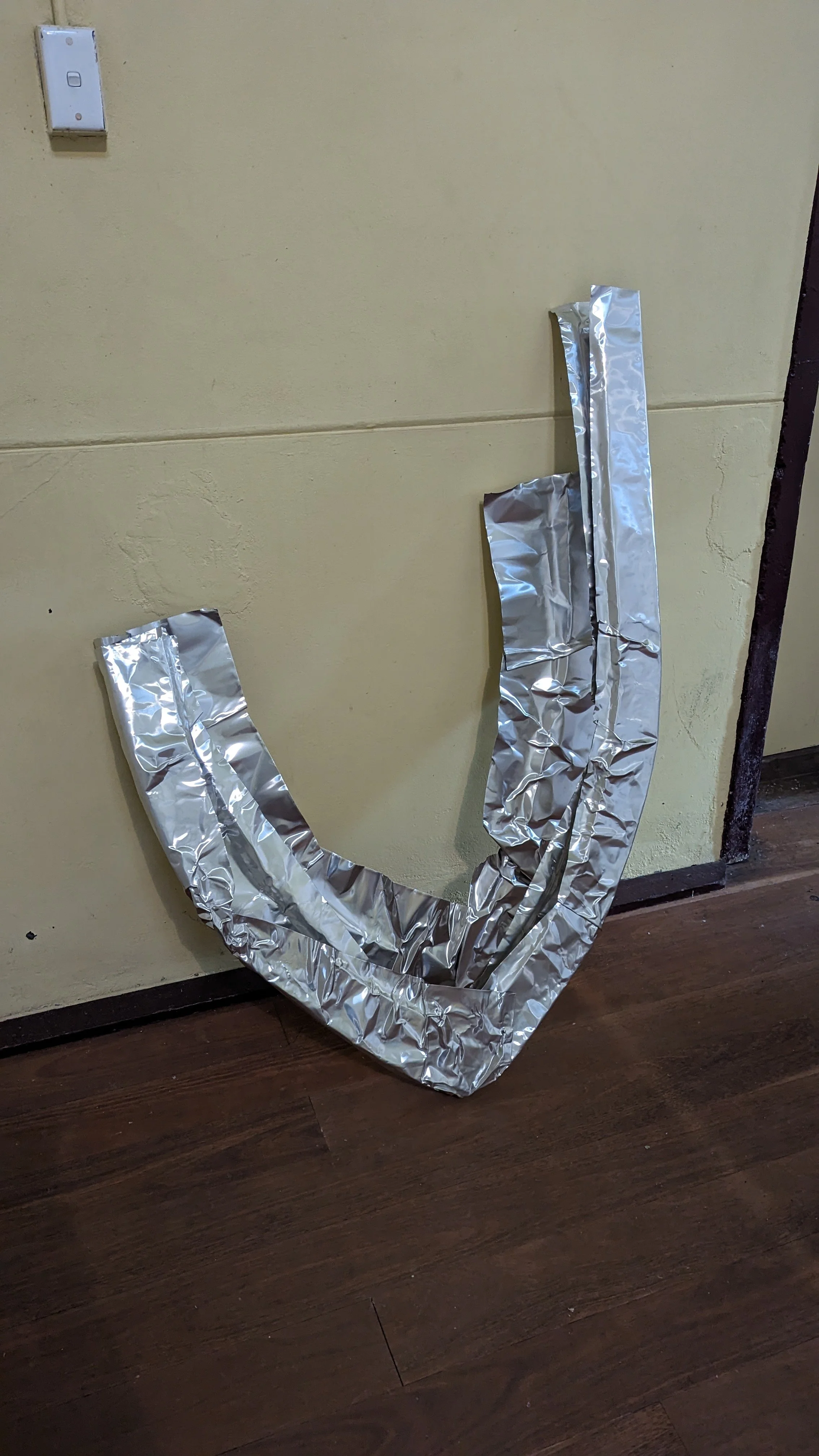 Aluminum foil shaped like a letter J leaning against a beige wall near a light switch and dark wooden floor.