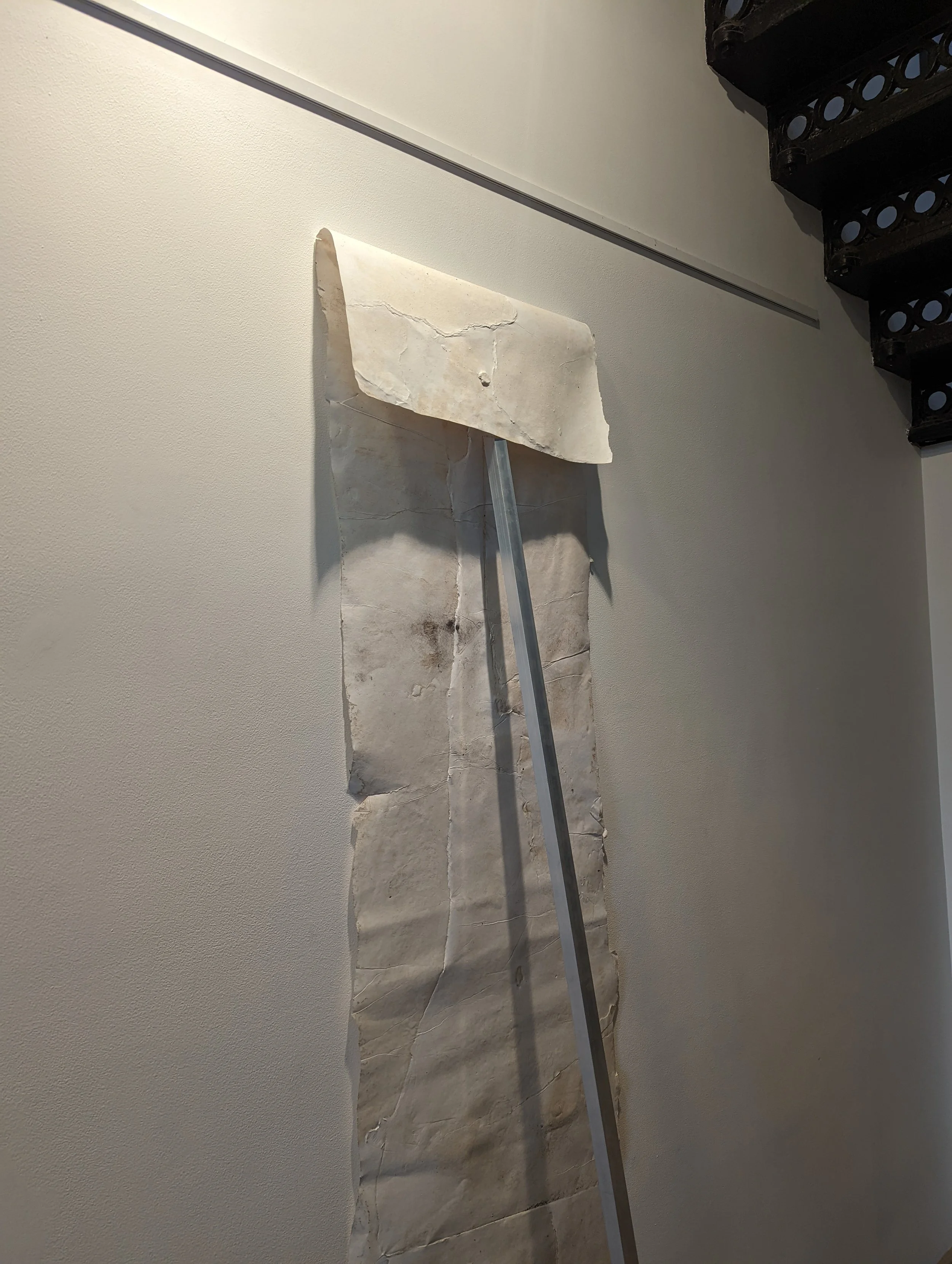 A marble or stone sculpture or structure leaning against a wall, with a pole or rod attached to it, and a dark ornate wooden shelf in the background.