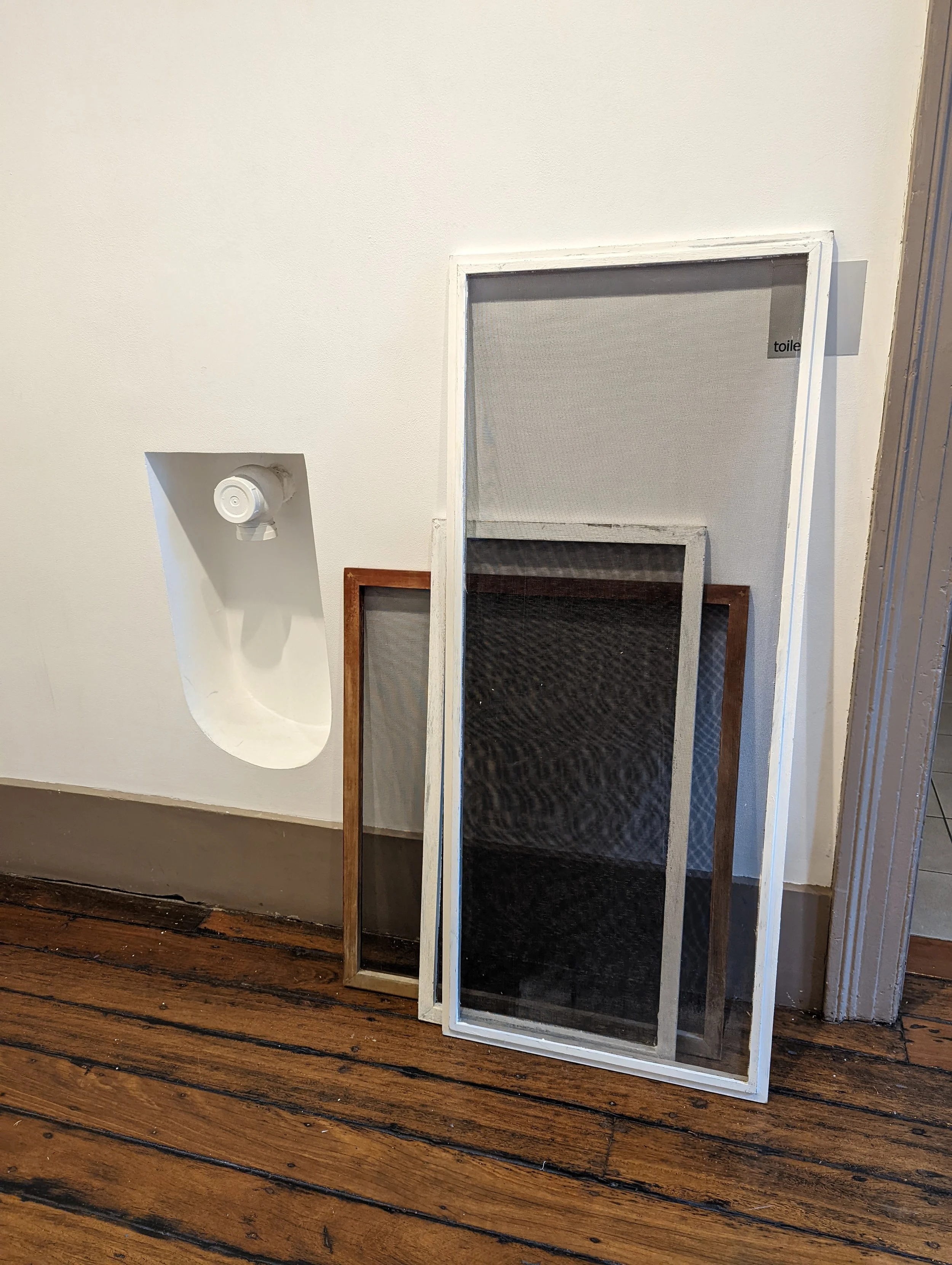 Three window screens leaning against a white wall, with a small section of a smoke detector on the ceiling and a wooden frame, on a wooden floor.