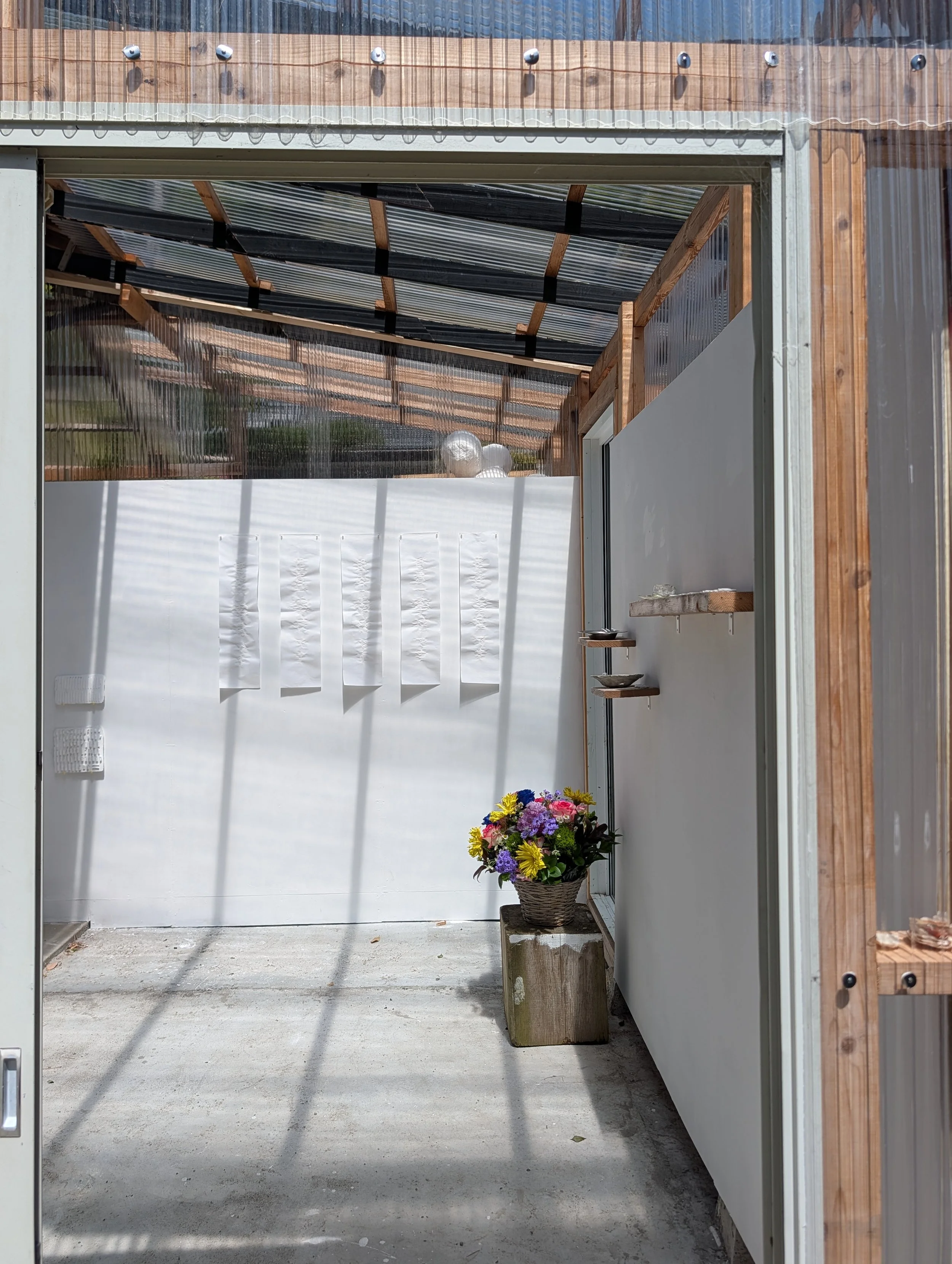 View through an open garage door showing a wooden planter with colorful flowers next to the wall indoors and a transparent curtain with sunlight outside.