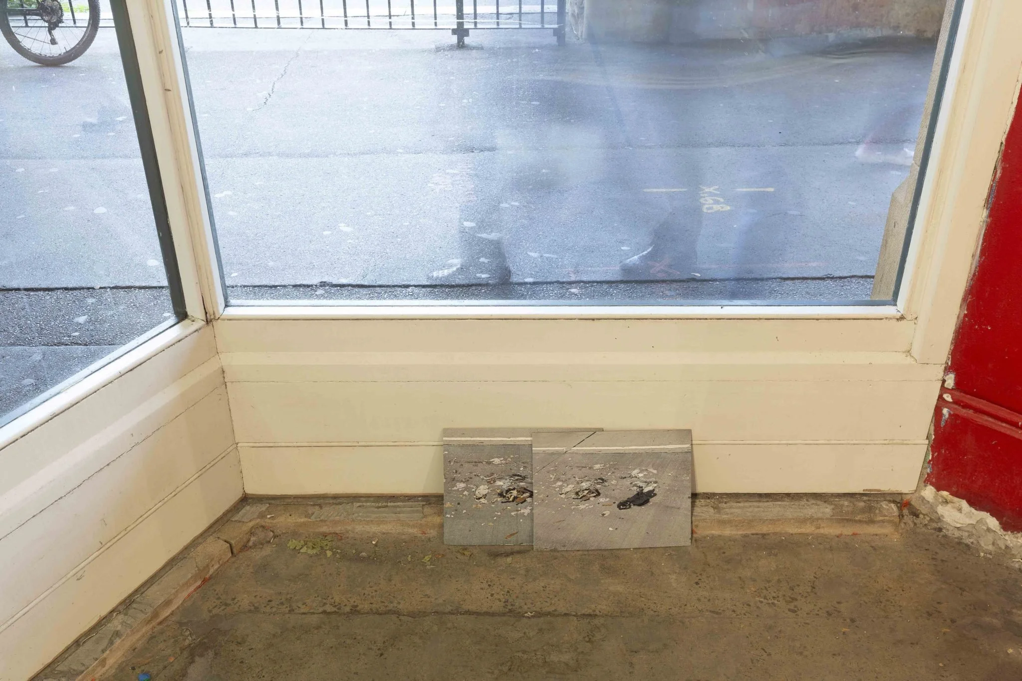 Interior view of a storefront entrance with a glass door, showing a sidewalk and street outside, with a bicycle parked nearby. The floor inside is unfinished with exposed concrete and debris.