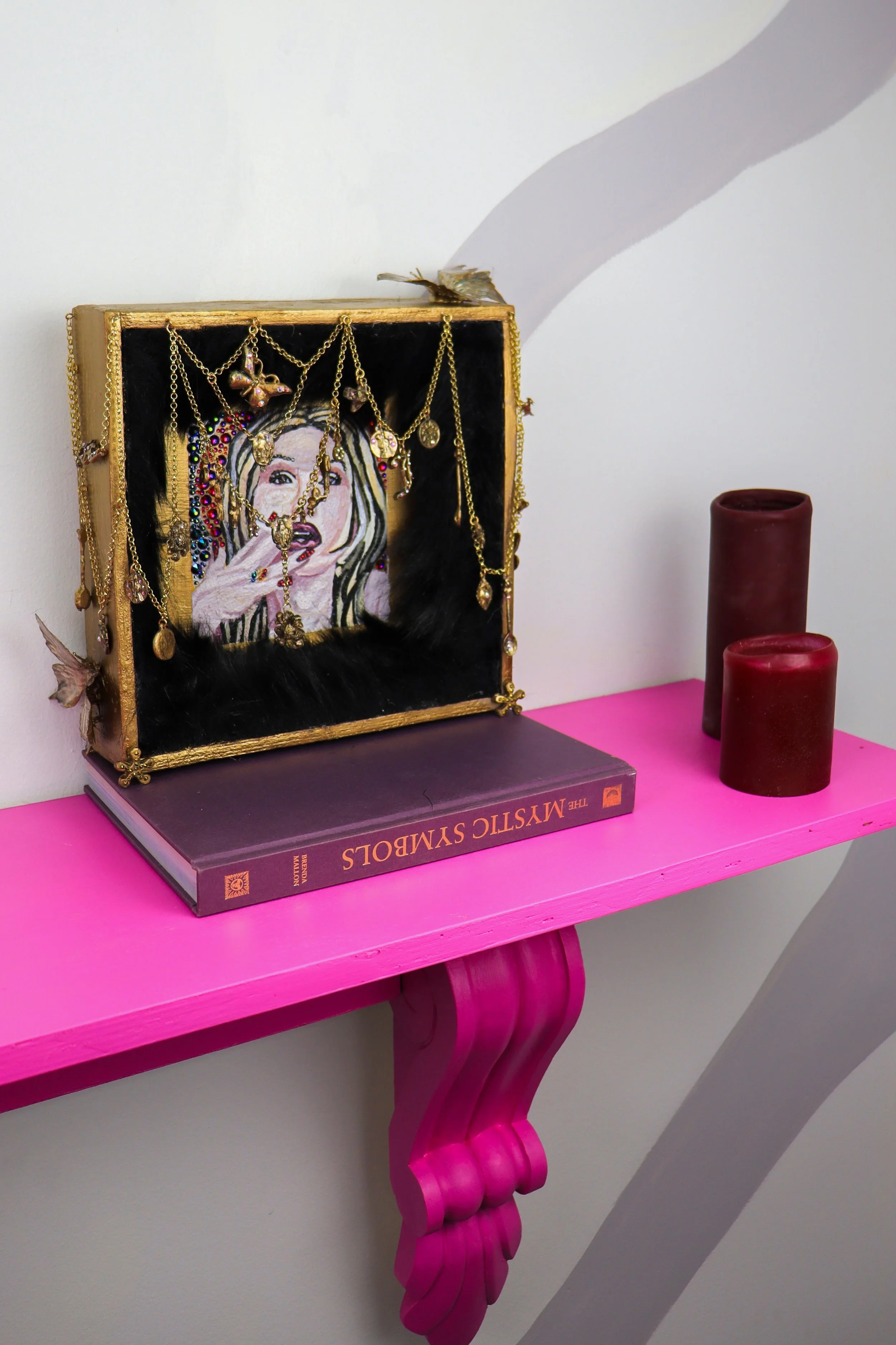 Decorative pink table with ornate curved leg, topped with a book and a pink wall shelf. On the shelf is a jewelry box with a portrait of a woman surrounded by necklaces, earrings, and other jewelry, along with two red candles.