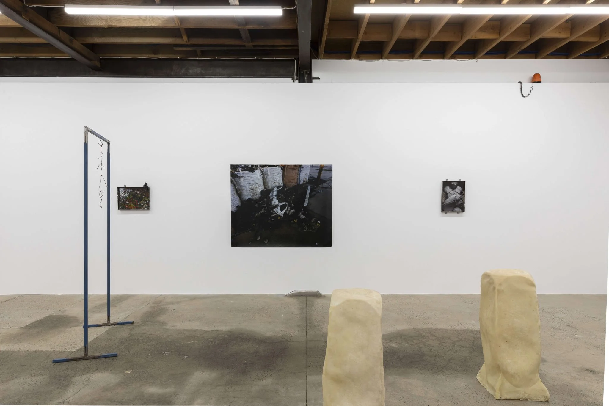 An art gallery exhibition featuring photographs on a white wall. There are two rocks in the foreground and a vertical sculpture with a string of lights on the left. The ceiling shows exposed wooden beams and fluorescent lighting.