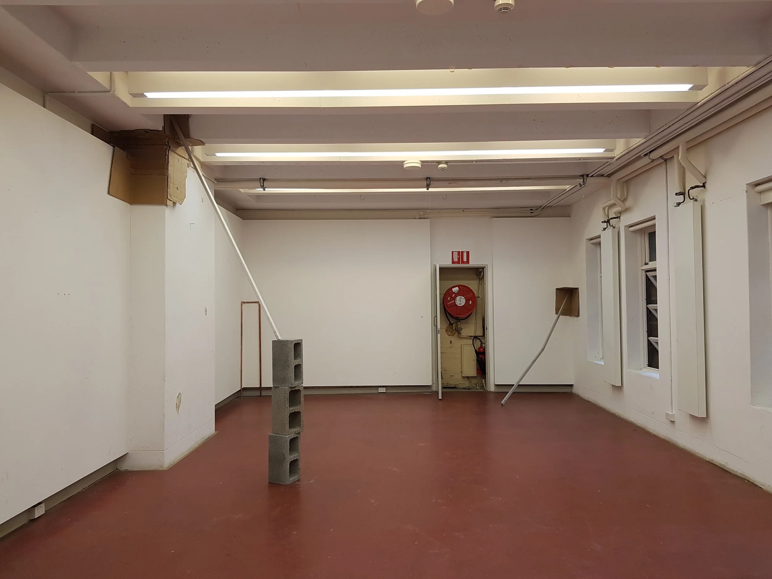 Empty room with white walls, fluorescent ceiling lights, and windows. There are construction materials like cinder blocks and a metal pole indicating ongoing renovation.